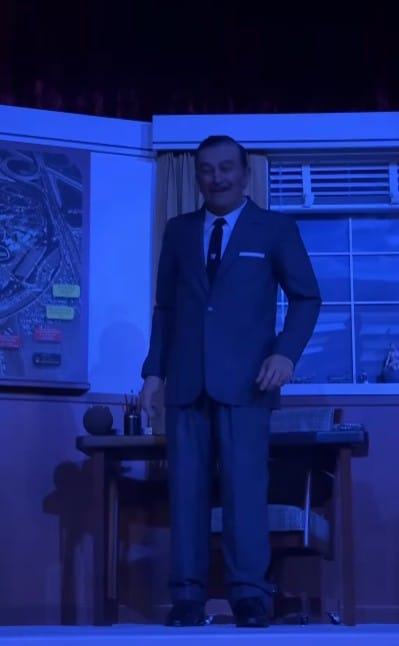 A man in a suit stands before a desk and large map, bathed in blue light, evoking classic Walt Disney innovation.
