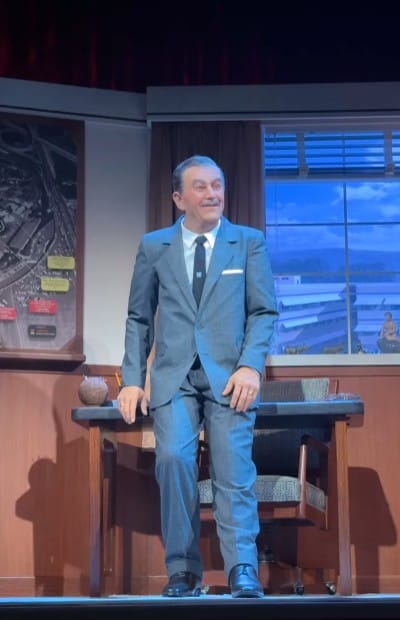 A man in a suit stands by an office desk, smiling, with a cityscape behind him like Walt Disney in his studio.
