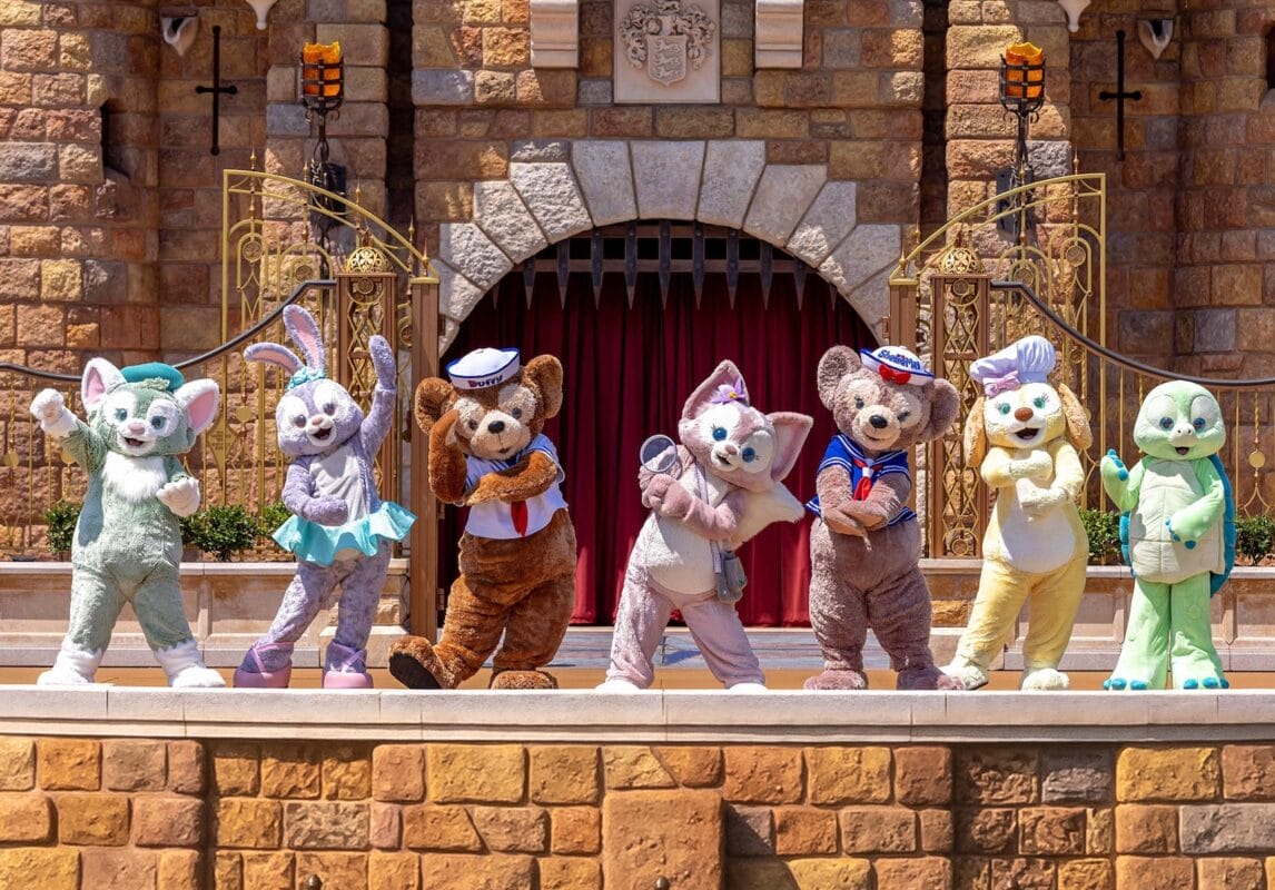 Six of Duffy's friends and Duffy the Disney Bear, pose on an outdoor stage before a stone castle backdrop.