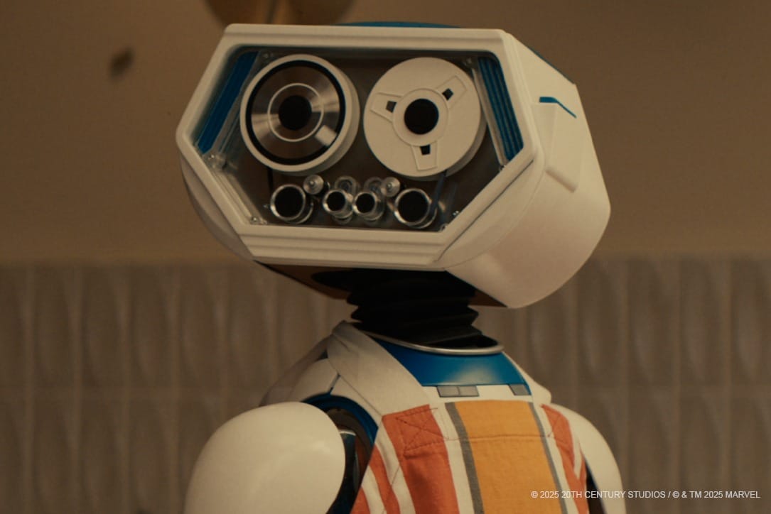 A close-up of a white and blue robot named H.E.R.B.I.E., with a hexagonal face and camera-like eyes, wearing an orange-striped apron.