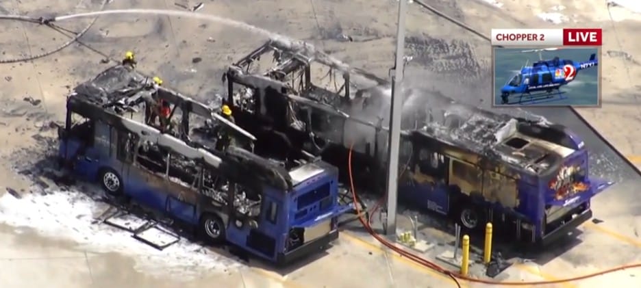 Two large buses, charred from a bus fire, sit in a lot as firefighters work below an Epic Universe news helicopter.