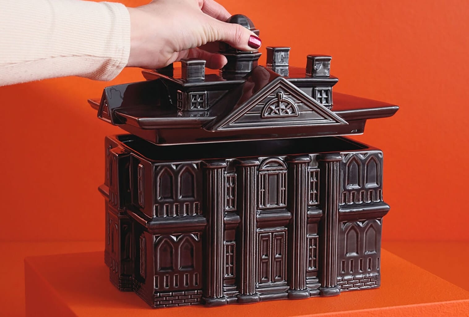A hand lifts the roof off a Haunted Mansion-inspired black ceramic cookie jar on an orange background.