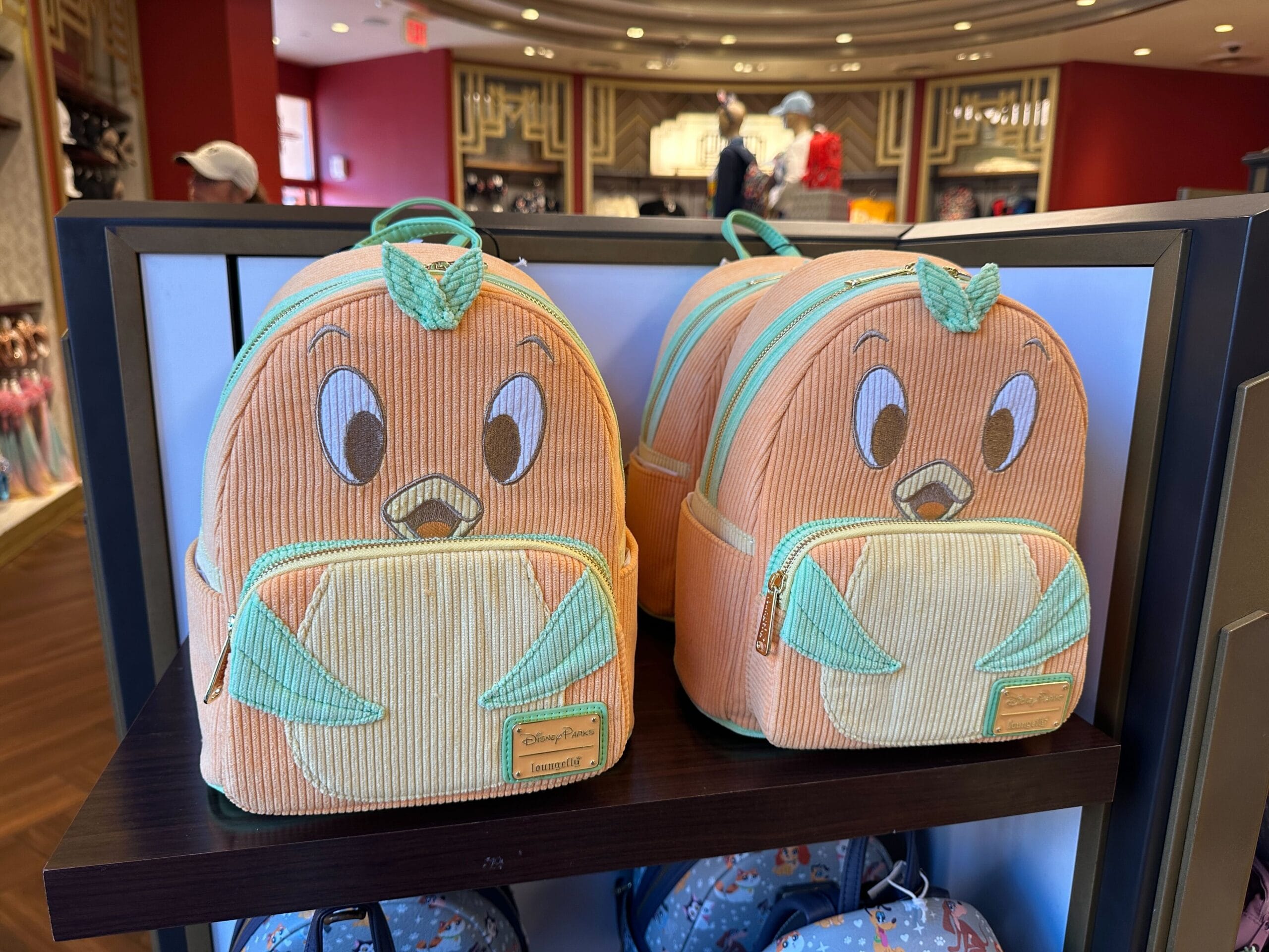 Two Loungefly Orange Bird-themed backpacks sit on a shelf in a brightly lit store.