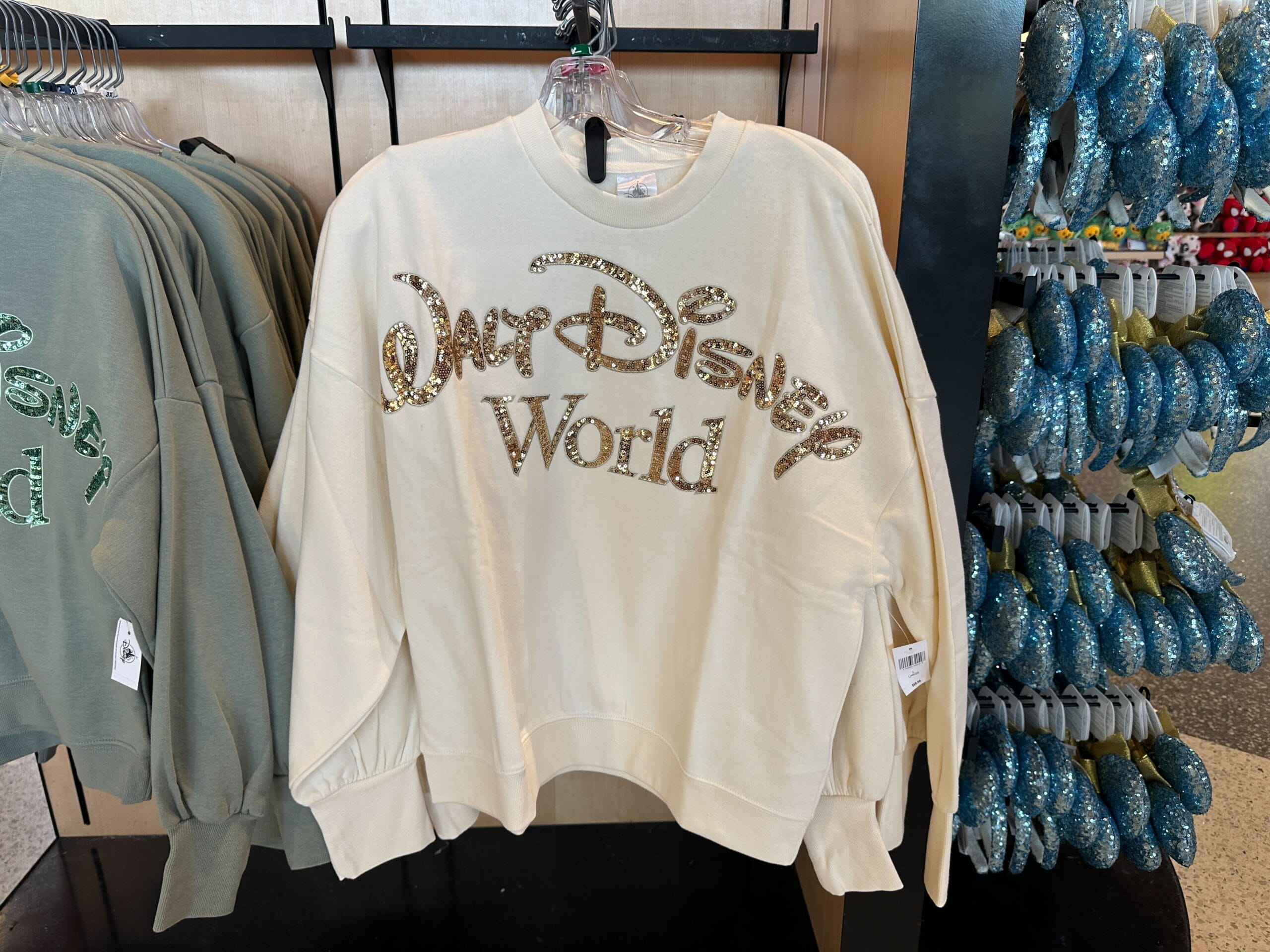 Cream sweatshirt with "WDW" in gold glitter letters, part of the new merch, hanging beside green sweatshirts.