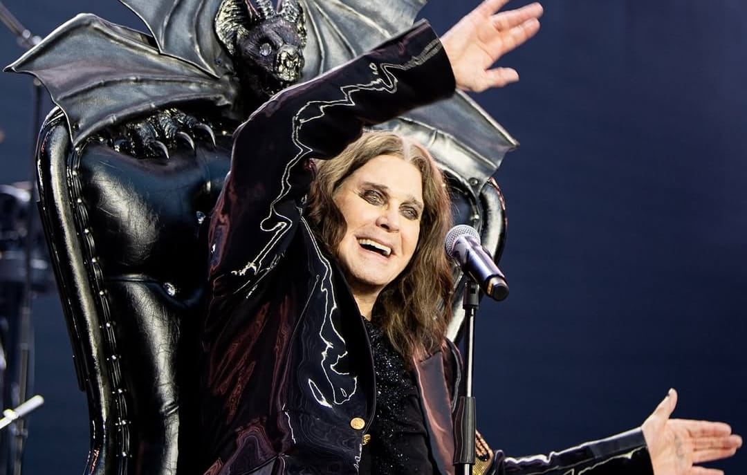 A long-haired performer in black sits on a gothic throne, smiling and waving—Ozzy Osbourne, Black Sabbath icon, at the mic.
