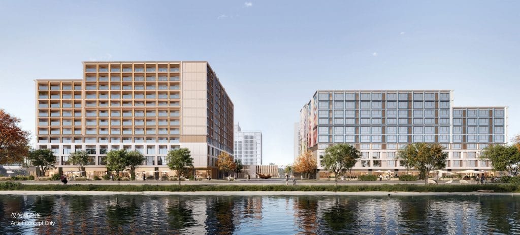 Two modern mid-rise buildings face a river, with trees and people along the waterfront under a clear sky.