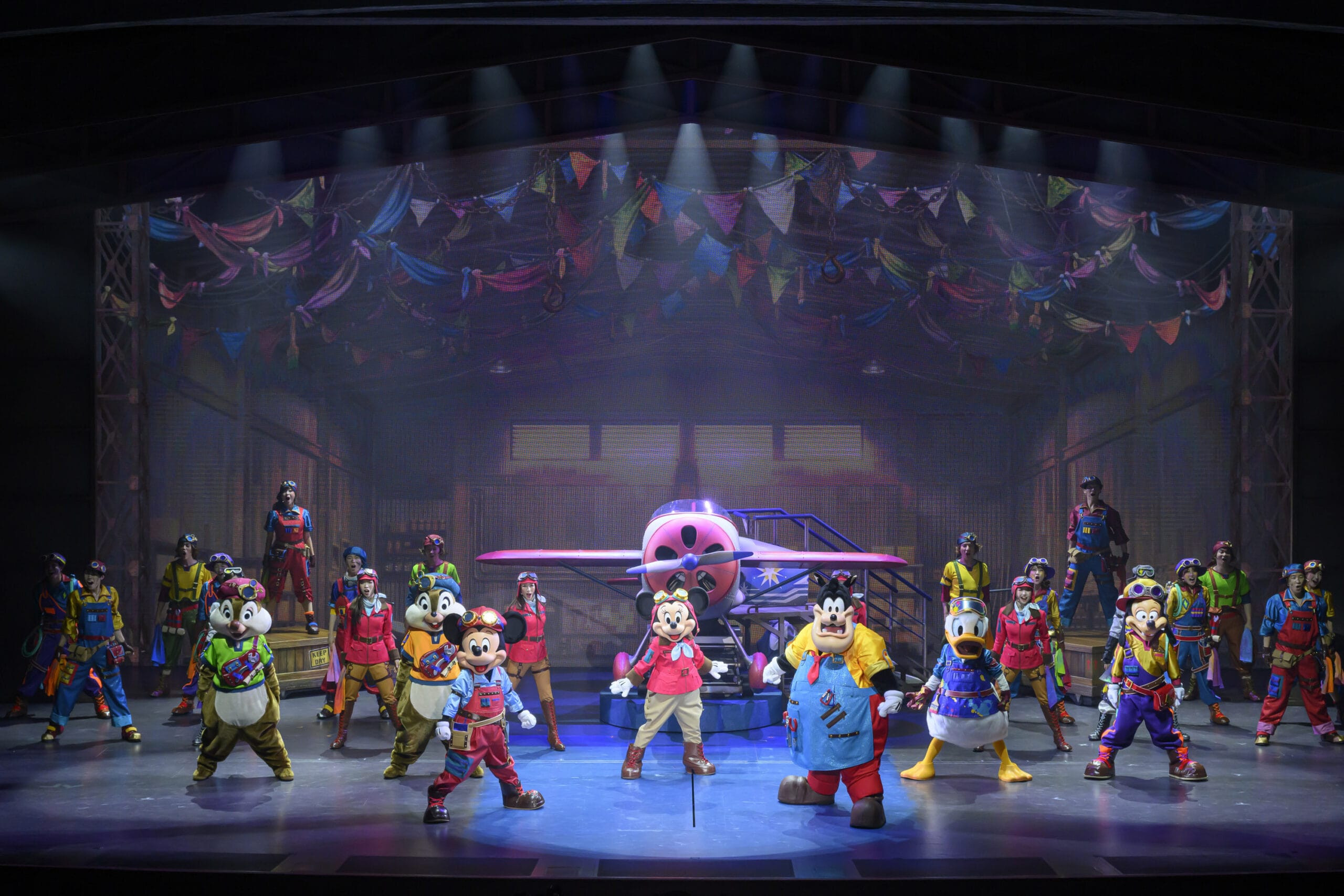 Performers in colorful costumes, where dreams take flight, stand on stage with Disney characters and a red airplane under bright lights.