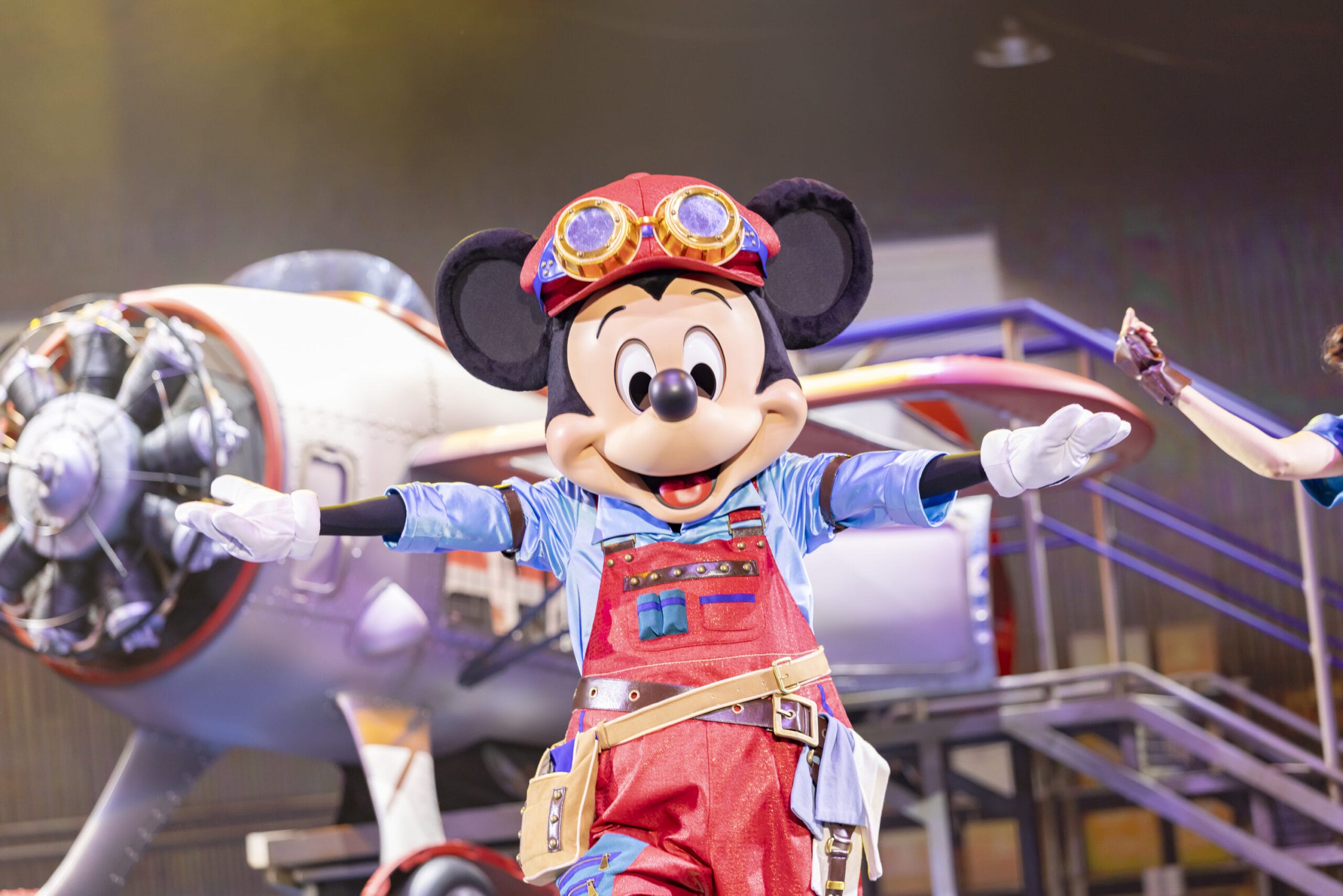 Mickey Mouse in red overalls and goggles poses as dreams take flight on stage in front of an airplane at Tokyo DisneySea.