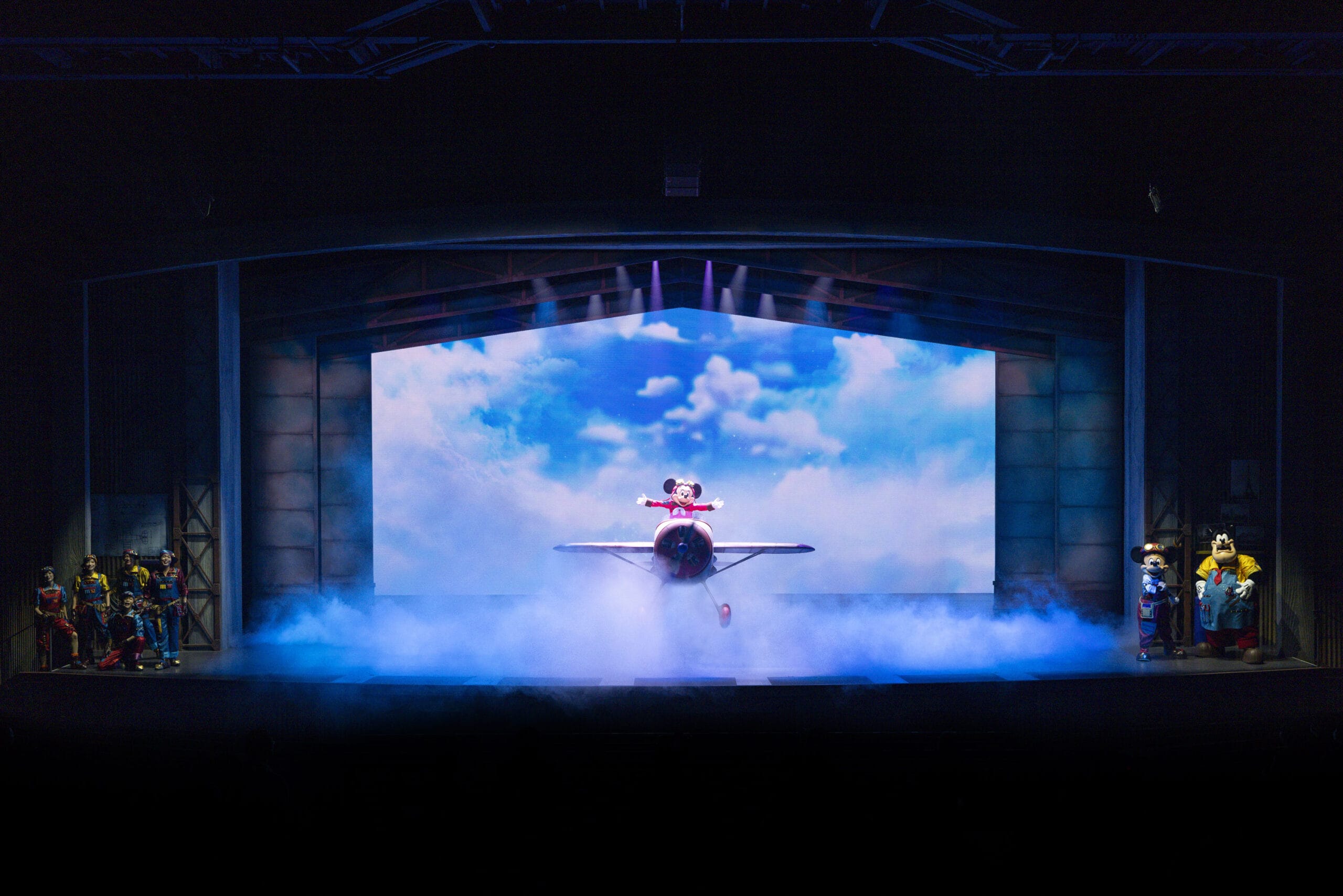 A stage at Tokyo DisneySea shows dreams take flight as a performer pilots a small plane amid smoke and actors.