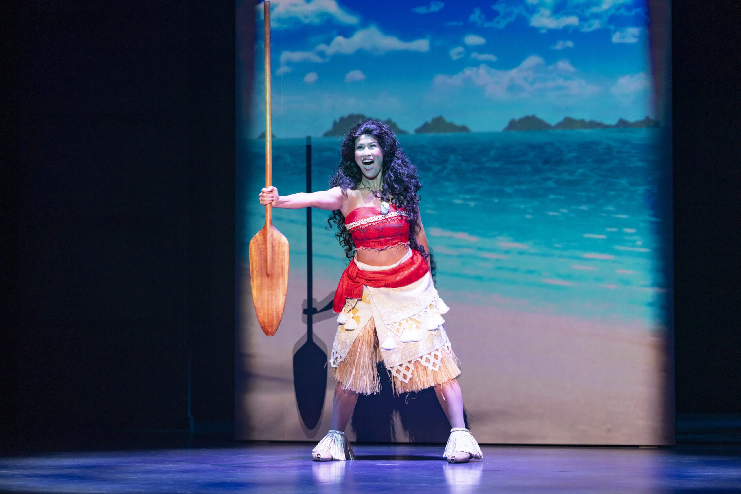 A woman in a tropical costume holds a paddle on stage, as dreams take flight at Tokyo DisneySea with an ocean backdrop.