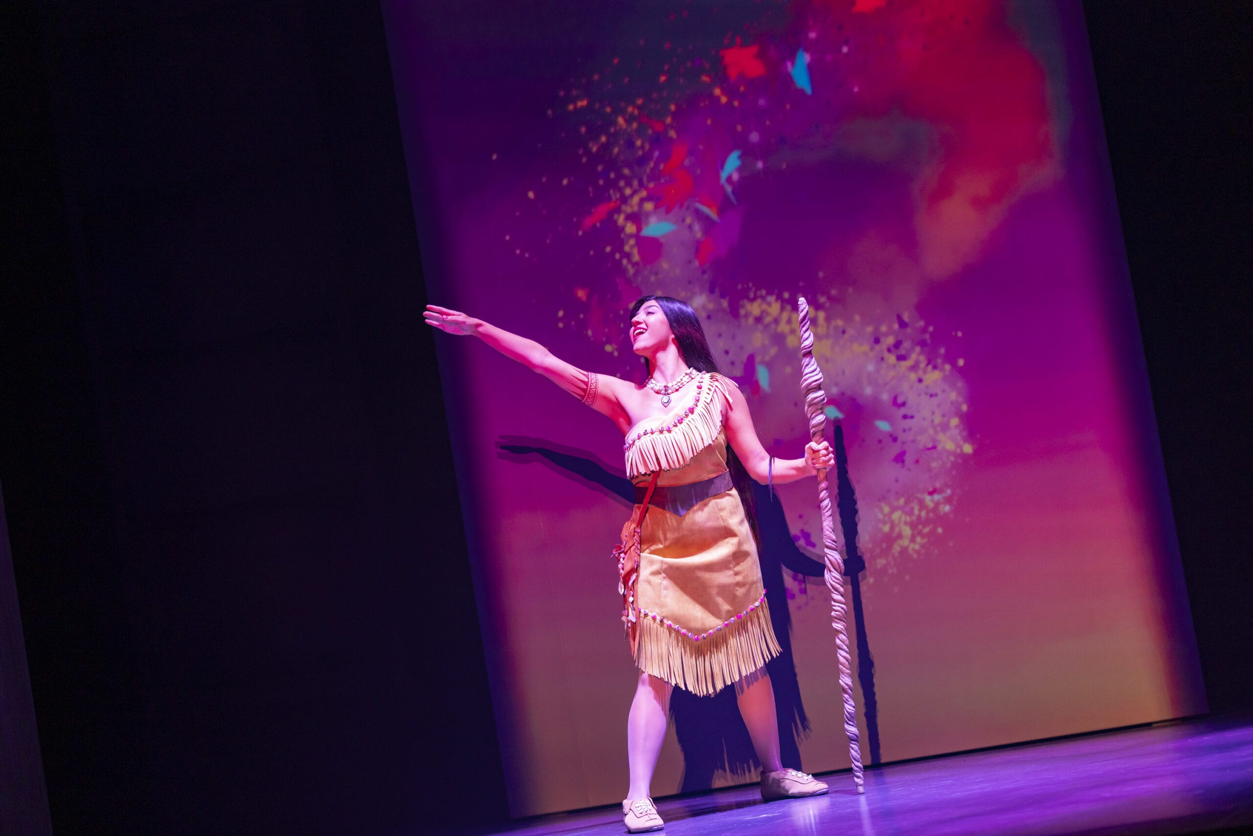 A woman in a tan fringe dress holds a staff, extending her arm on the colorful, lit stage at Tokyo DisneySea.