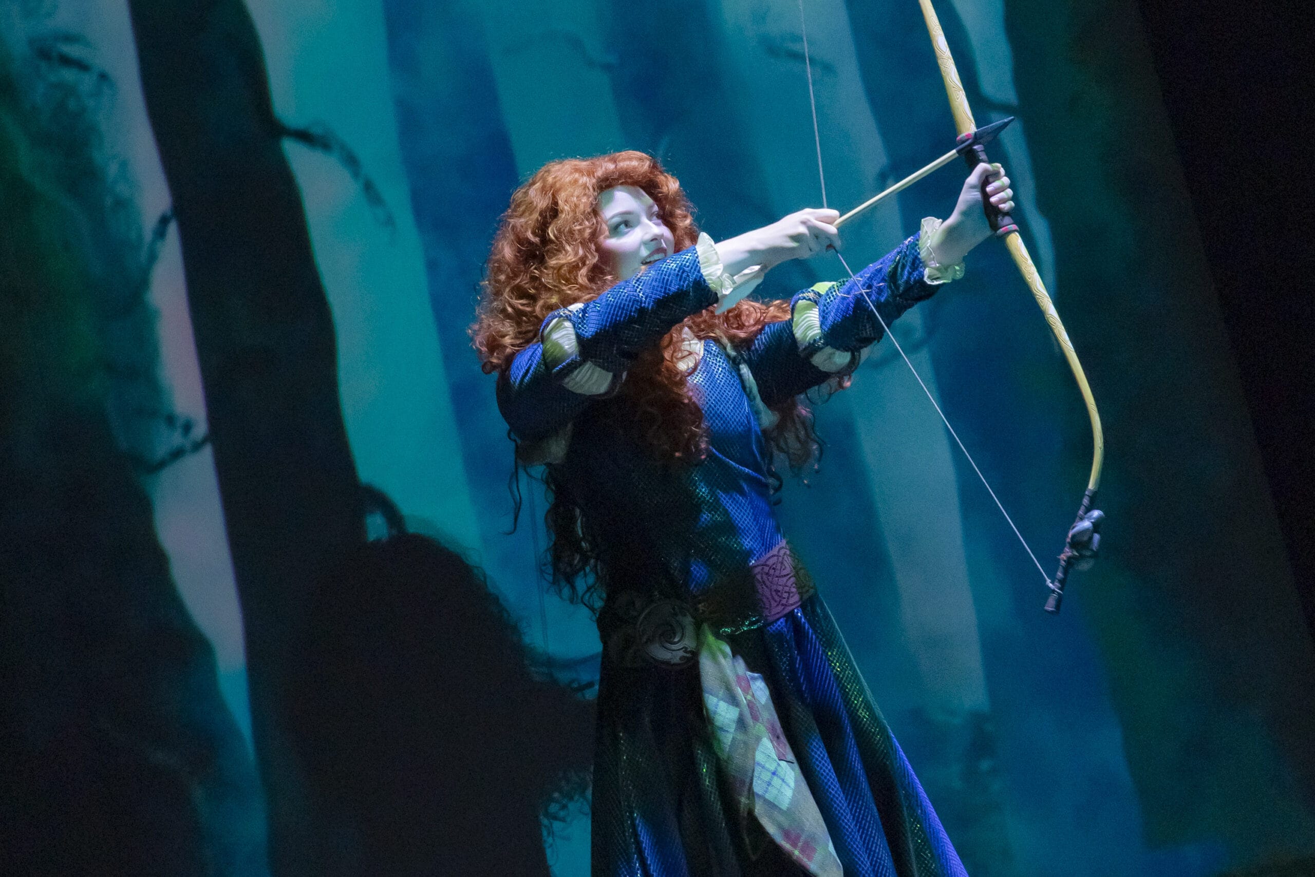 A woman with curly red hair aims her bow in a forest, wearing a blue dress—where dreams take flight at Tokyo DisneySea.