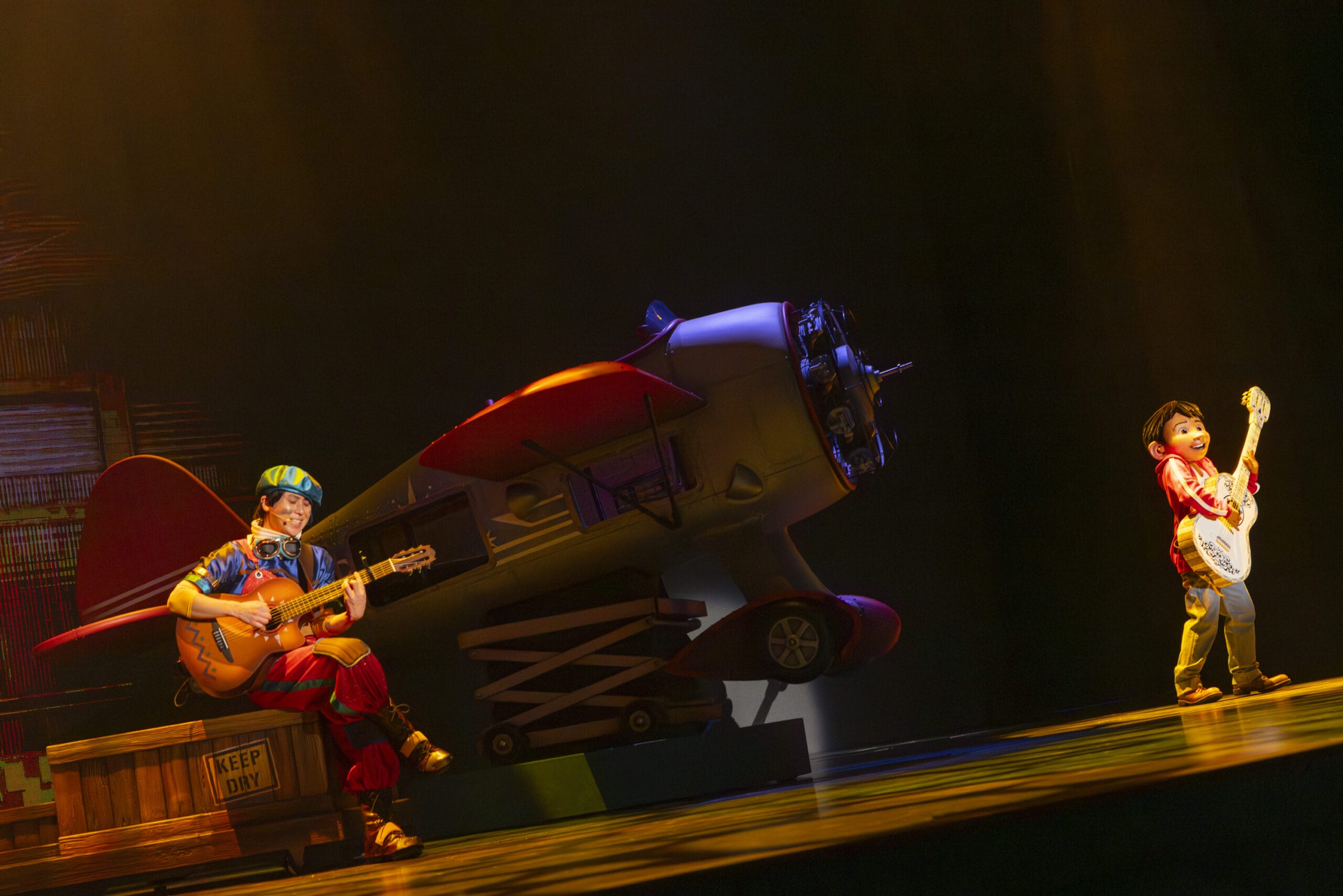 Two performers play guitars on stage at Tokyo DisneySea, as dreams take flight amid a colorful prop airplane and dramatic lighting.
