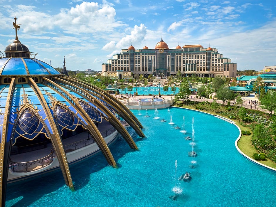 A grand hotel and domed building at Epic Universe overlook a turquoise pool with fountains under a blue sky.