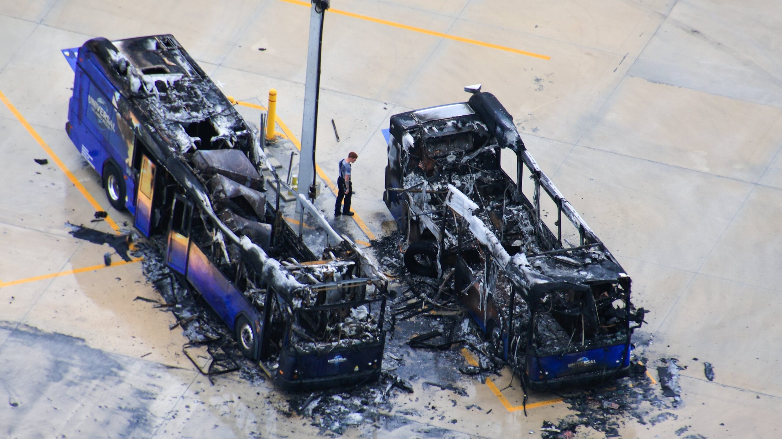 Three burnt-out electric buses with severe fire damage; a fireman stands nearby in the parking lot looking at the Universal bus fire damage.