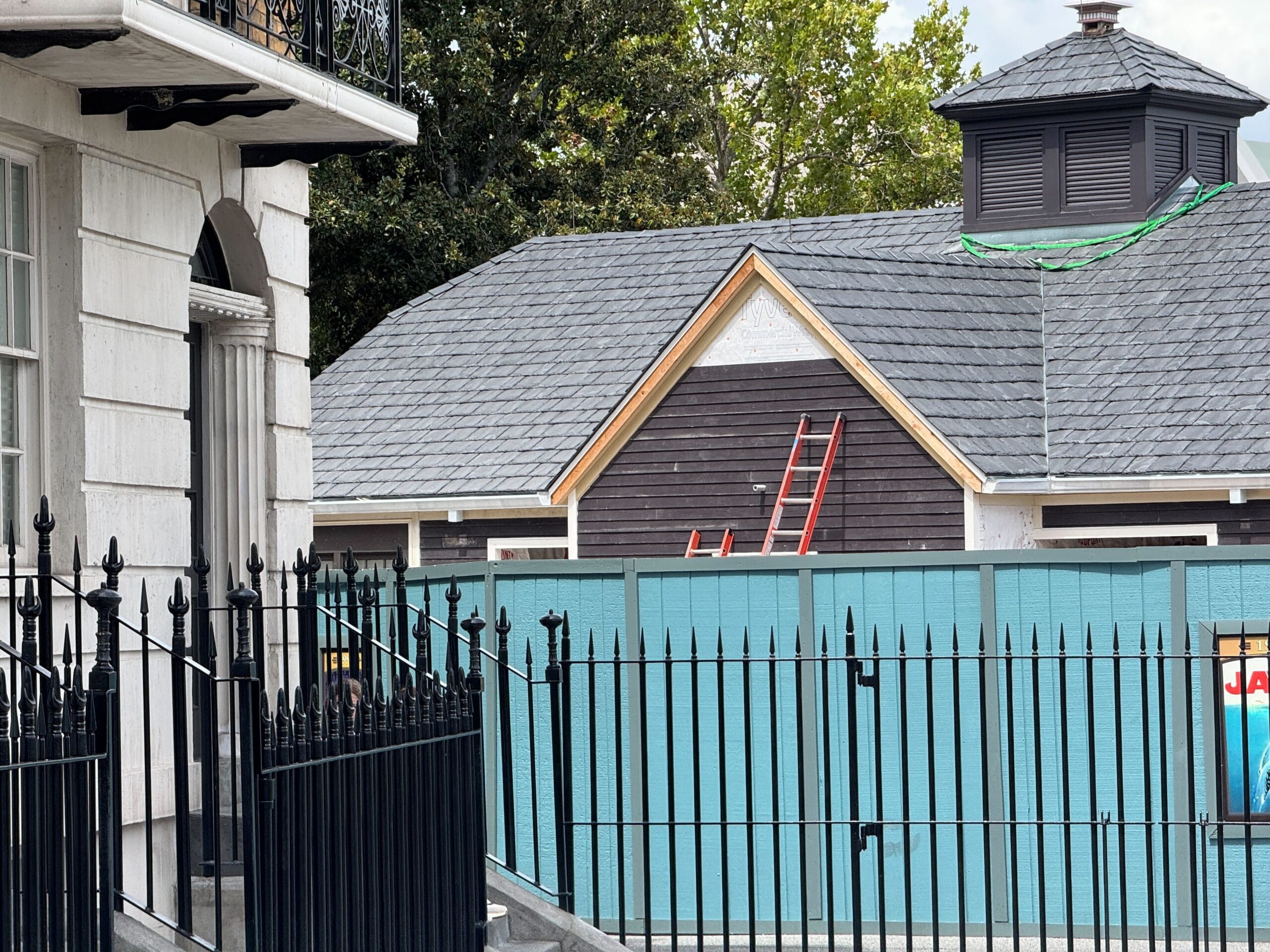 A Jaws bathroom building with a gray shingle roof, red ladder, and blue construction fence in front, next to a black iron gate.