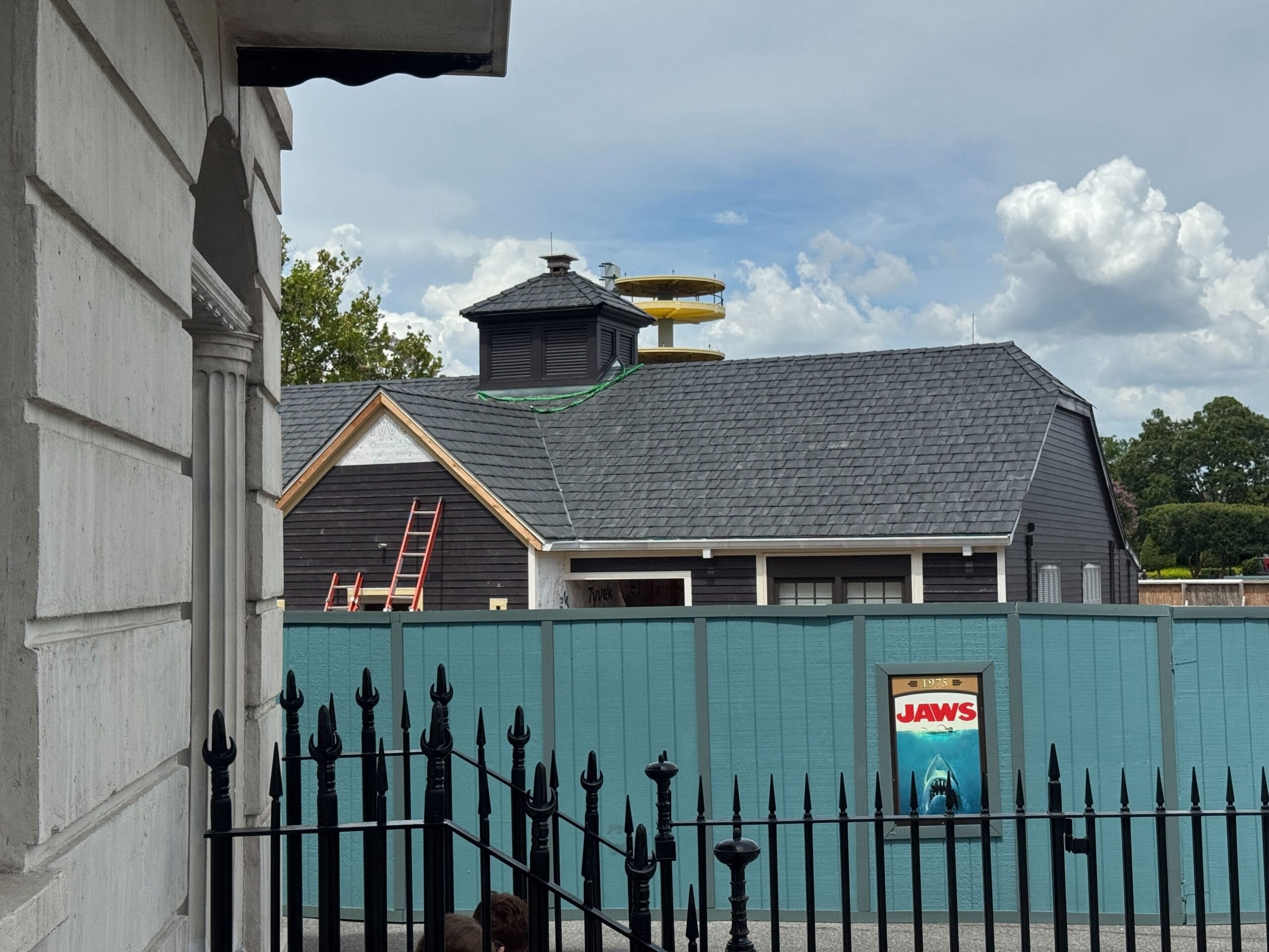 A bathroom building under construction behind a blue fence with a JAWS sign; a yellow slide is visible on the roof.