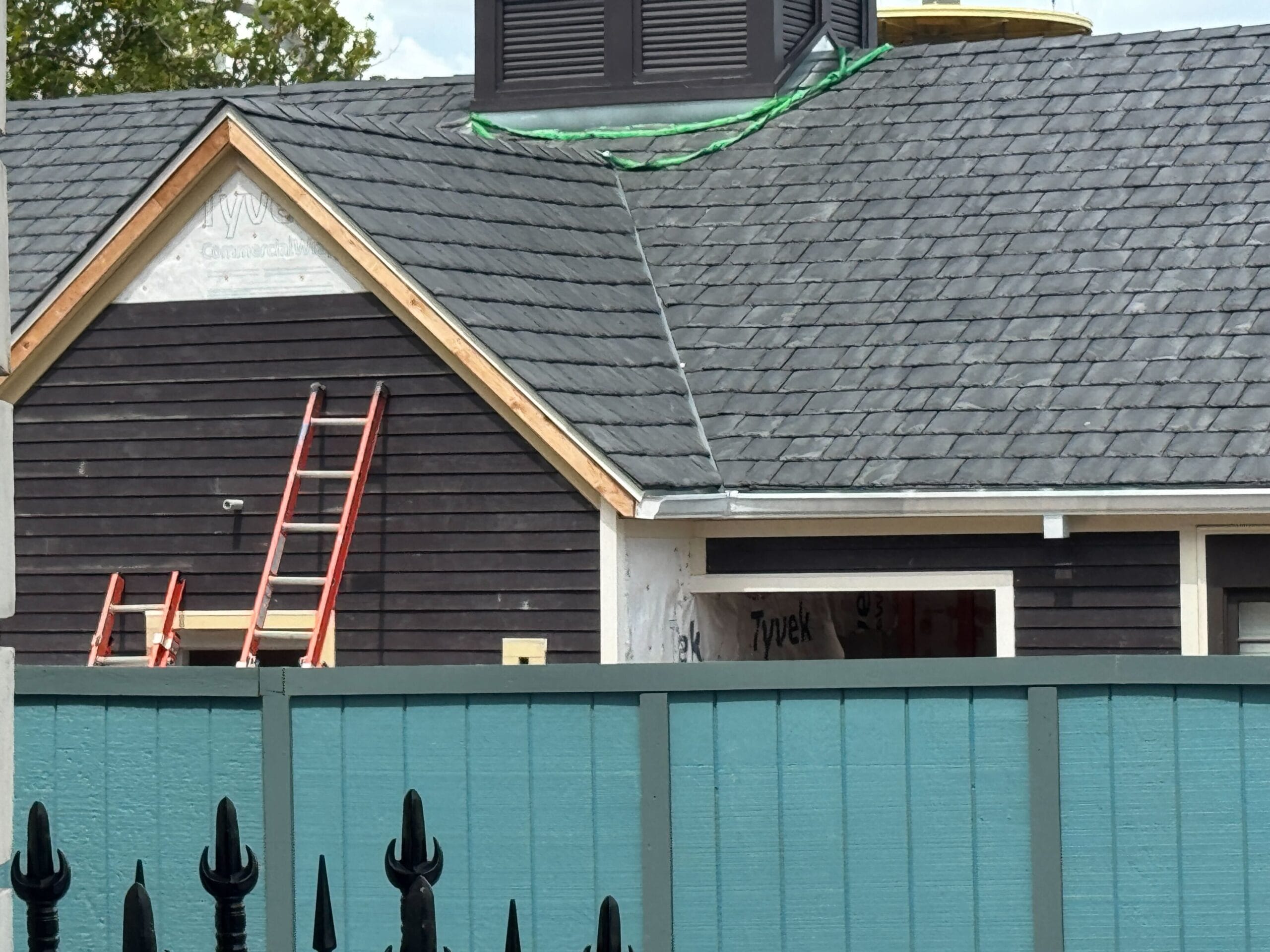 The Jaws bathroom under construction with ladders, dark siding, gray roof, and a blue fence in front.