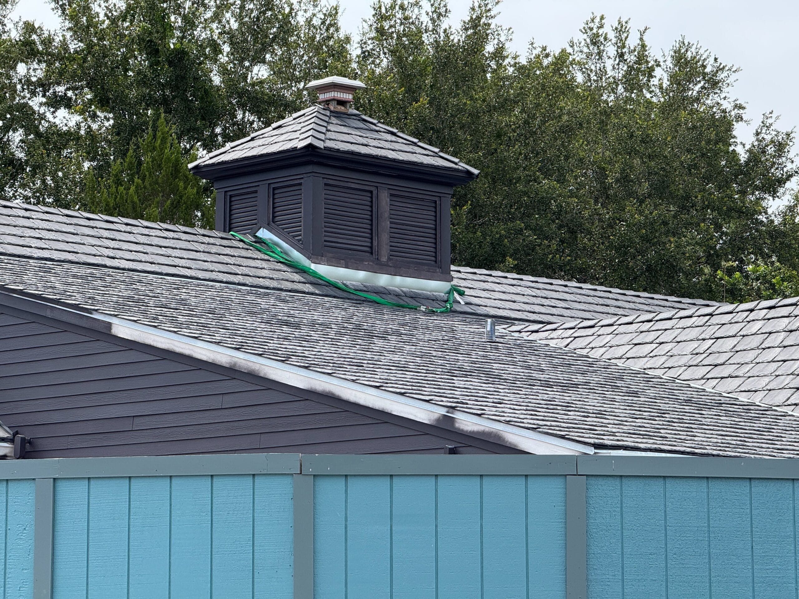 Gray shingled roof with a small cupola and a green strap, behind a turquoise wooden fence and trees in the background.