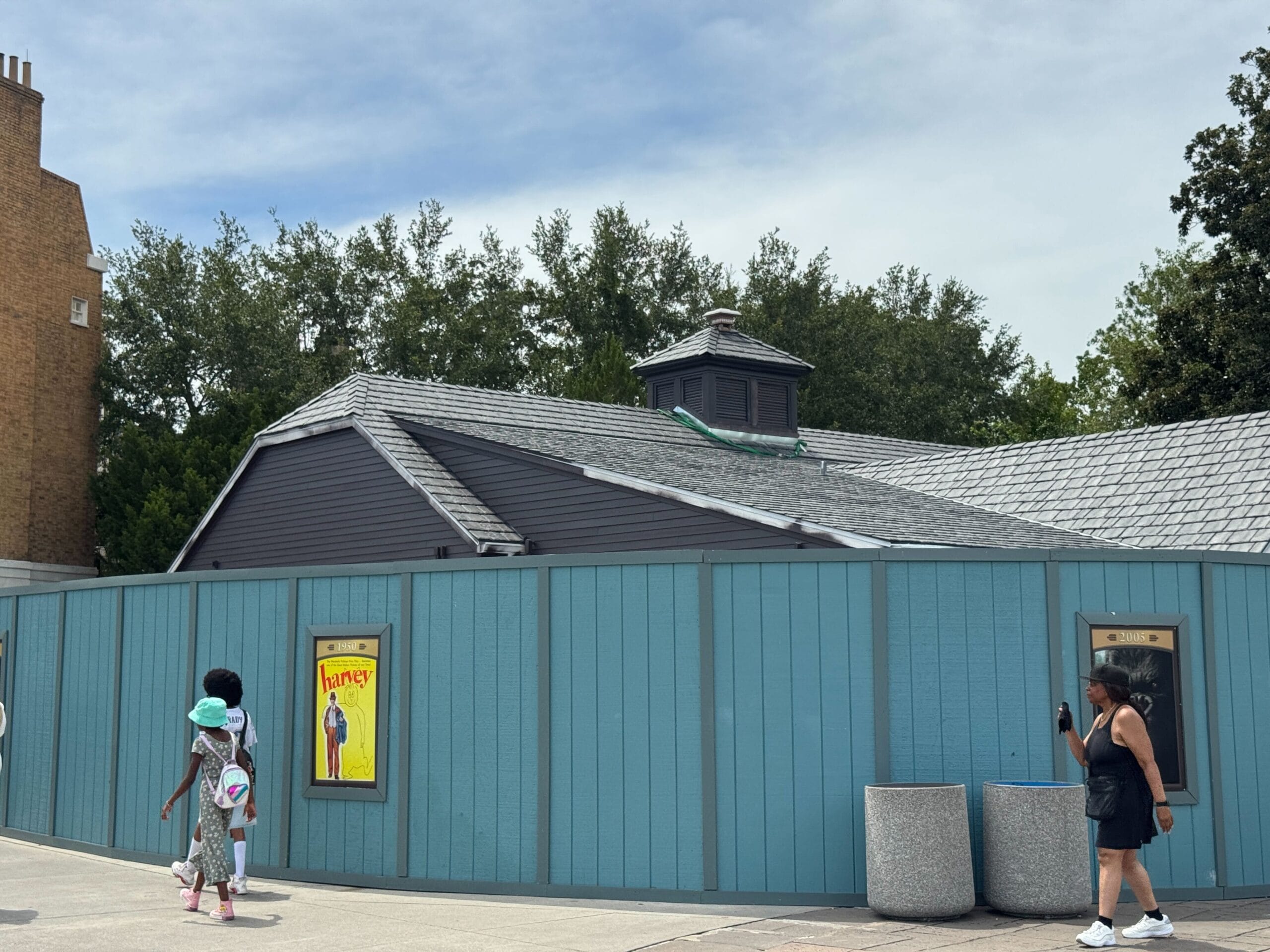 A blue construction wall surrounds a bathroom building in Universal Studios Florida; people walk by and two posters are displayed on the wall.