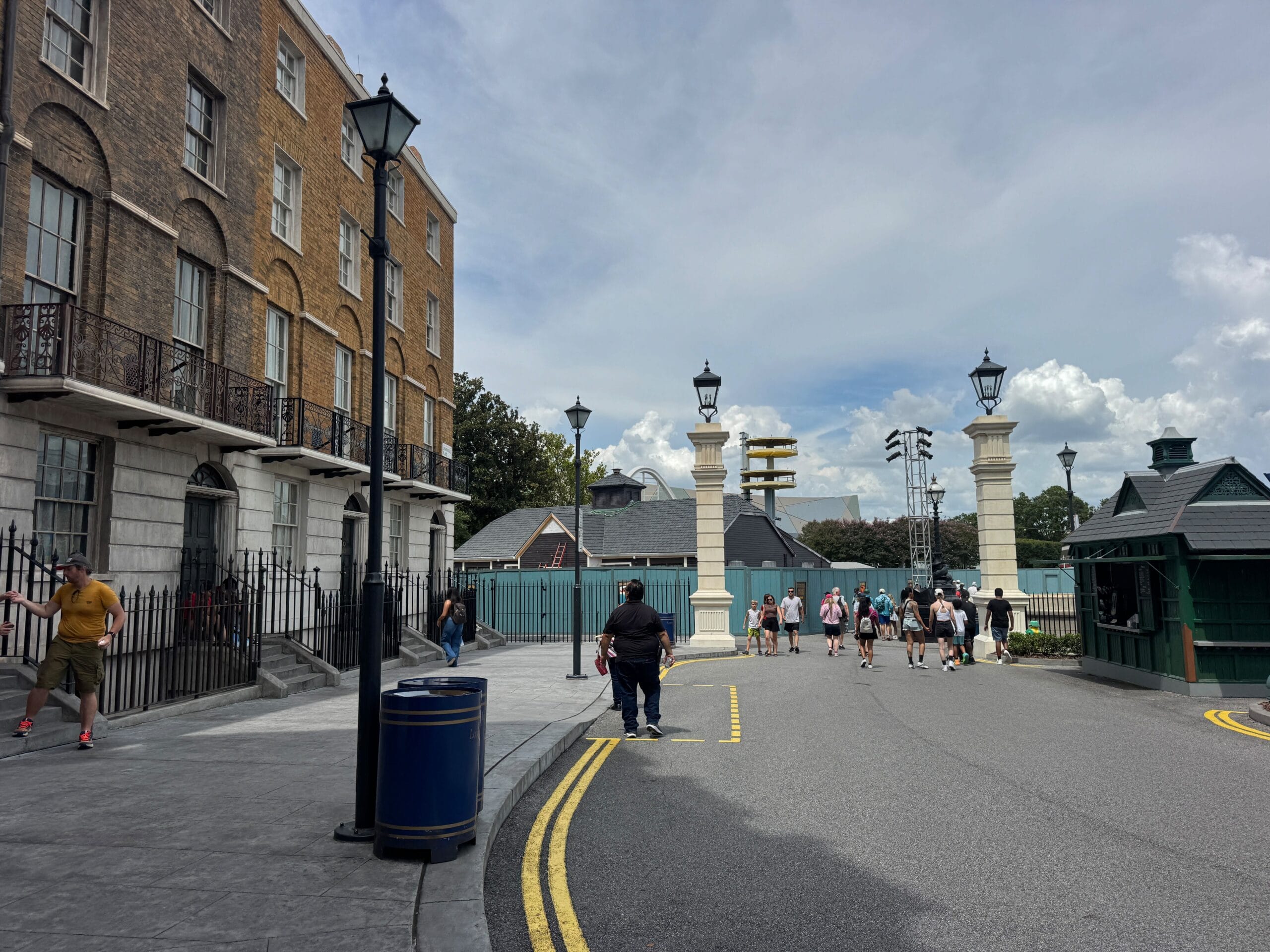 People walk near Grimmauld Palace and lamp posts on a curved street under a partly cloudy sky in Universal Studios Florida.