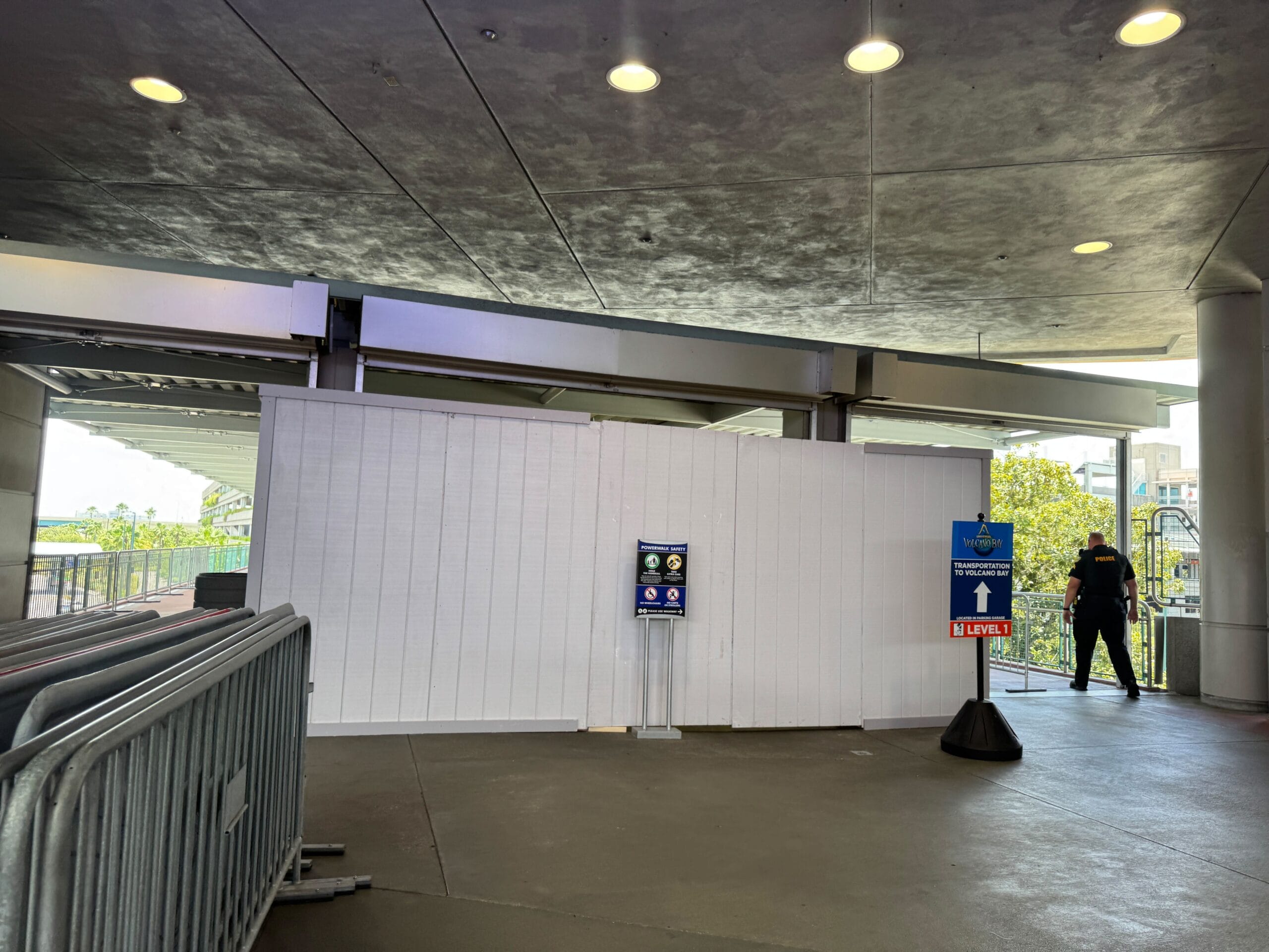 A white construction wall blocks a walkway; a security guard stands nearby, and signs give directions.