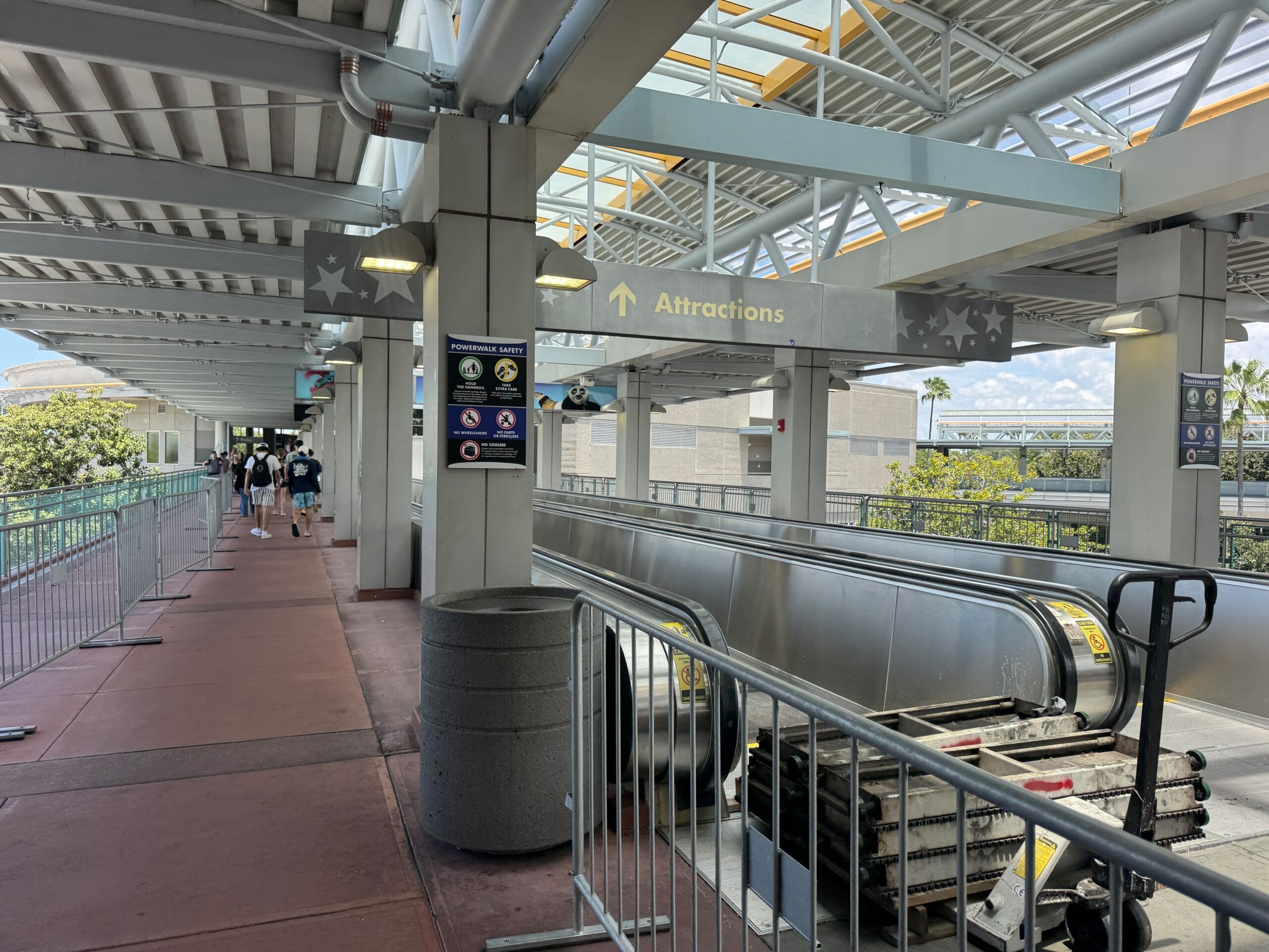 Outdoor walkway with people, overhead "Attractions" sign, escalator, and metal railings under a covered structure.