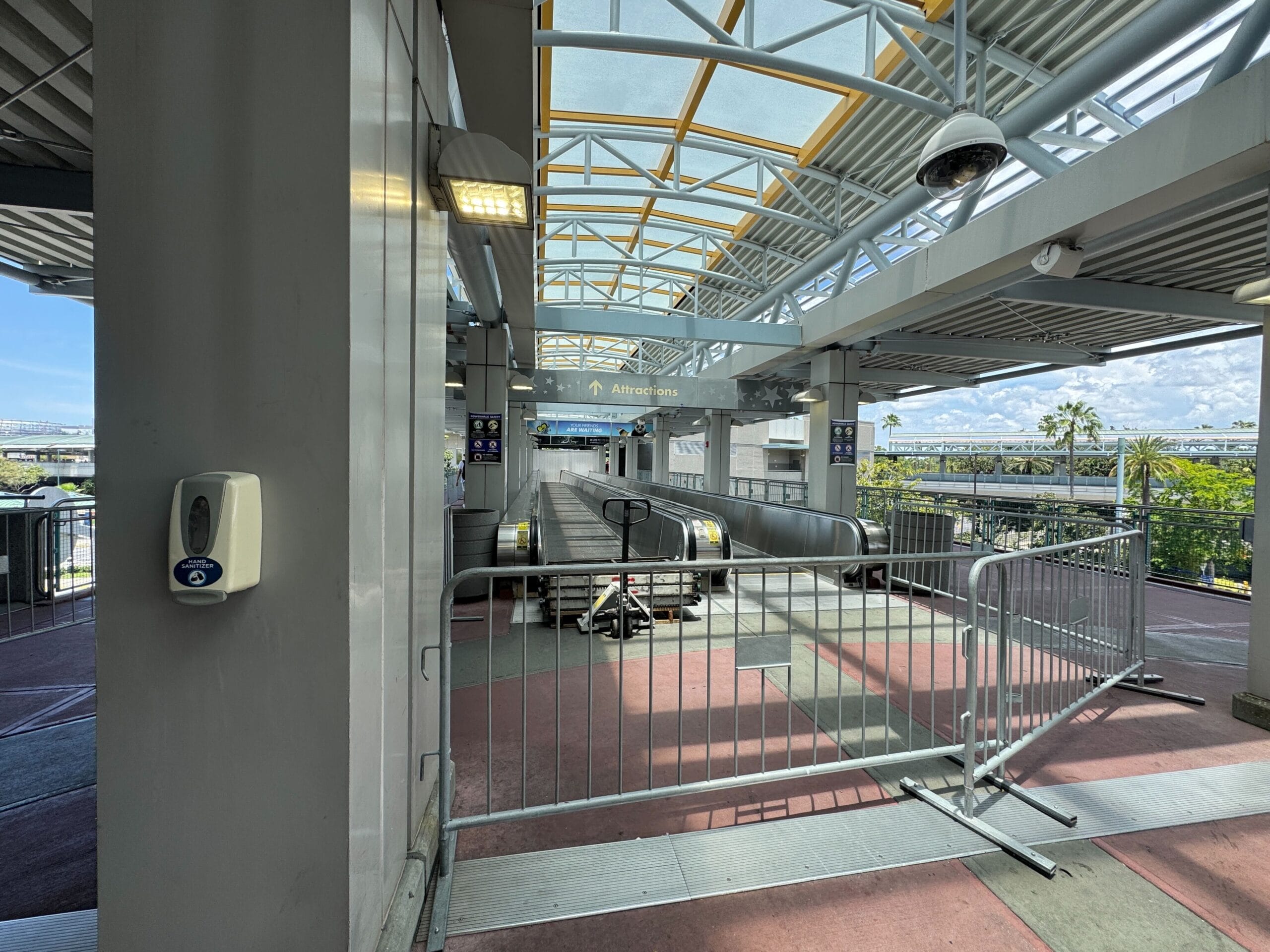 Outdoor moving walkway entrance blocked by metal barricades under a covered walkway, with a hand sanitizer dispenser on a column.