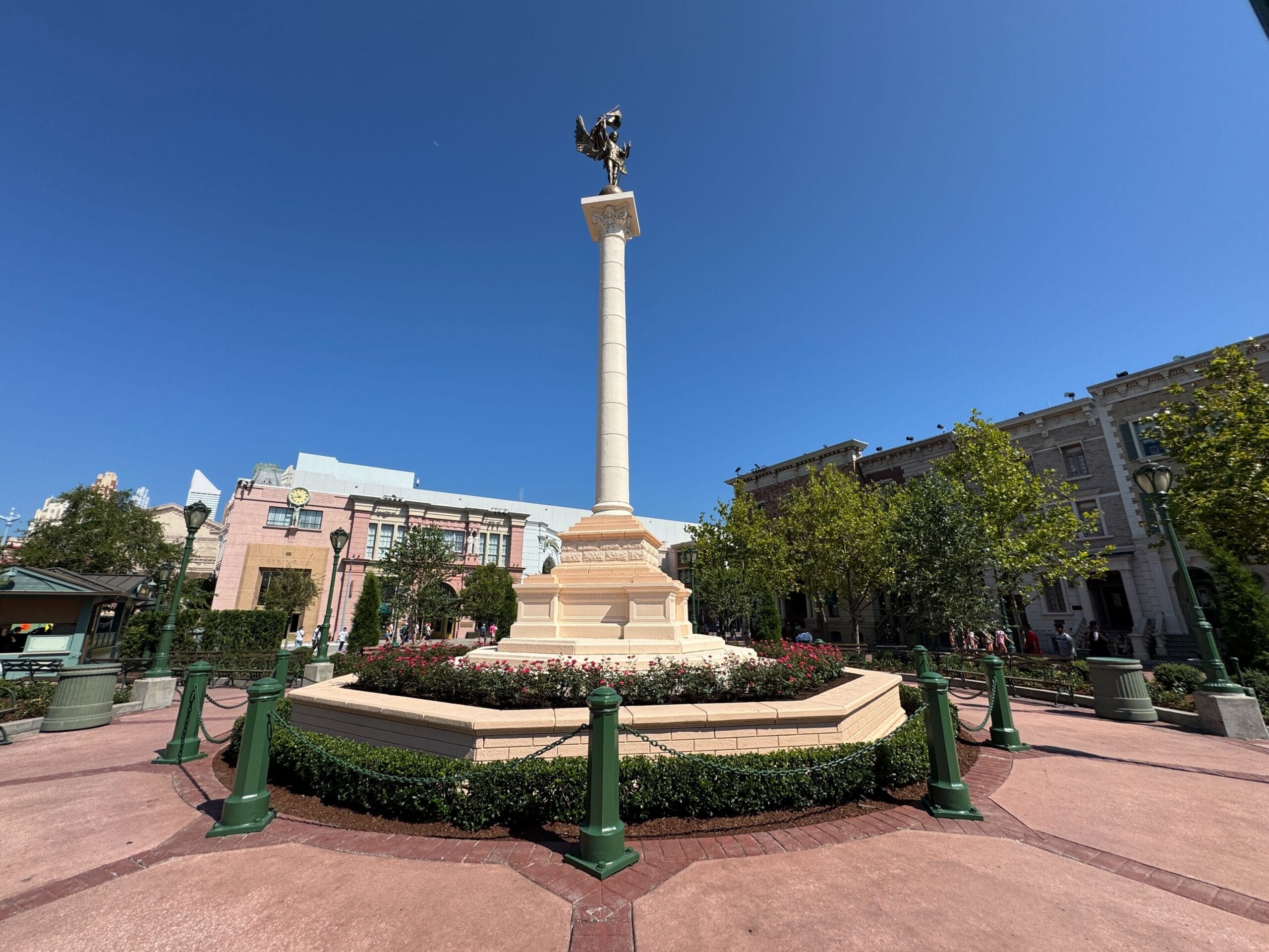 A tall column with a statue on top stands in a circular garden surrounded by buildings under a clear blue sky.