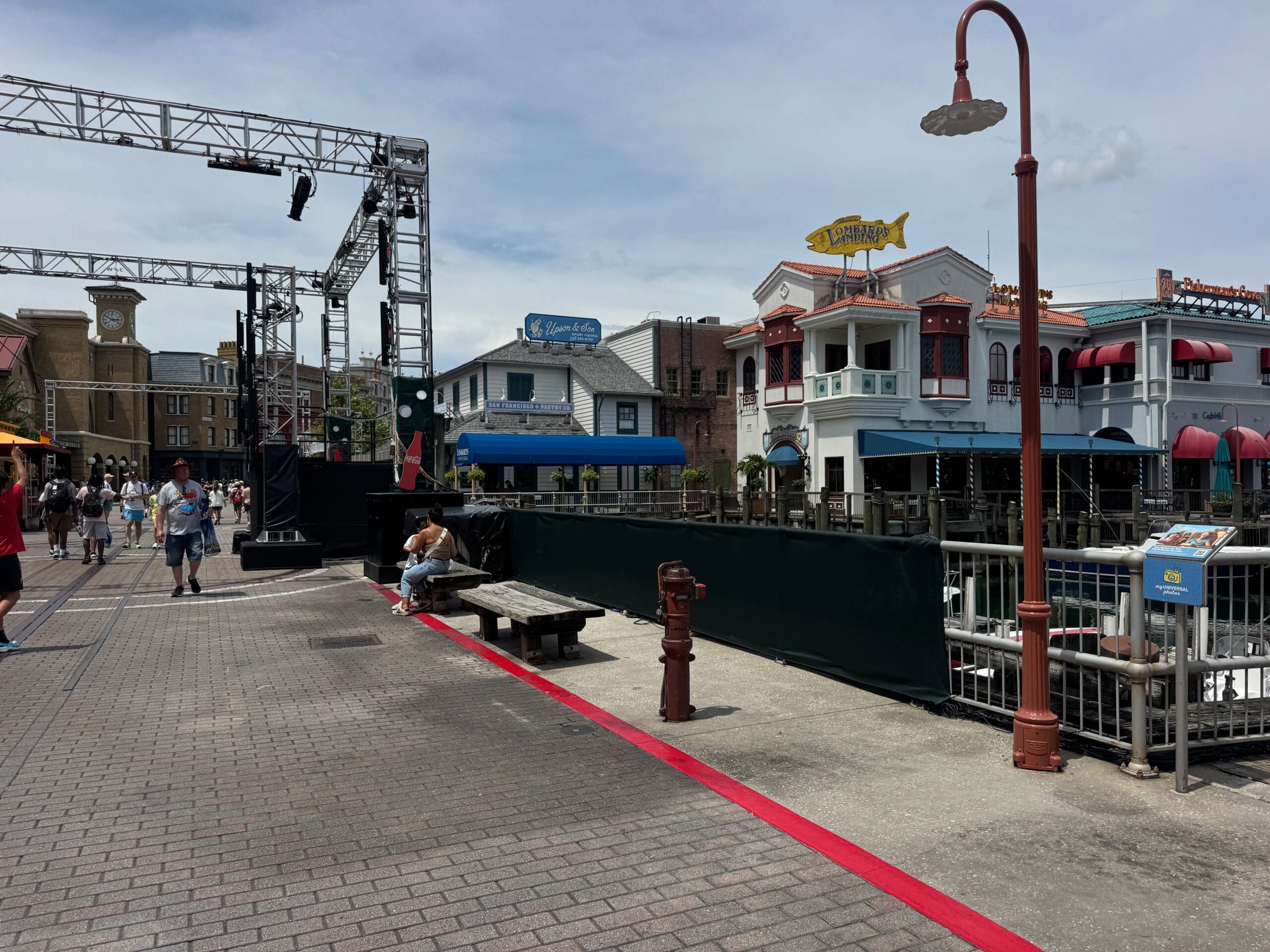 Theme park street during Halloween Horror Nights, with visitors, colorful buildings, scaffolding, and a red line under cloudy skies.