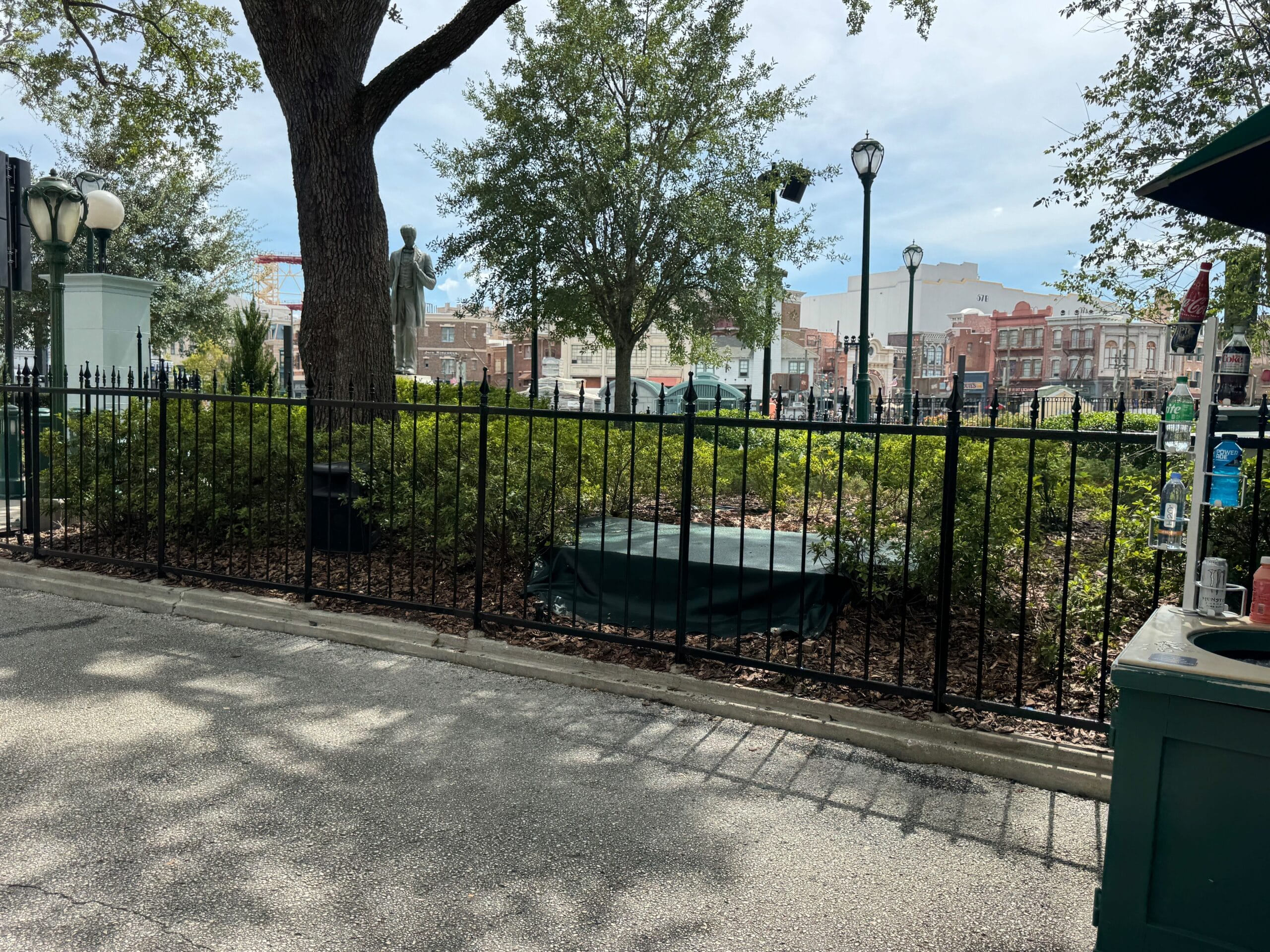Black iron fence encloses a shrub area, hinting at a Scare Zone for Halloween Horror Nights with trees and covered statue.
