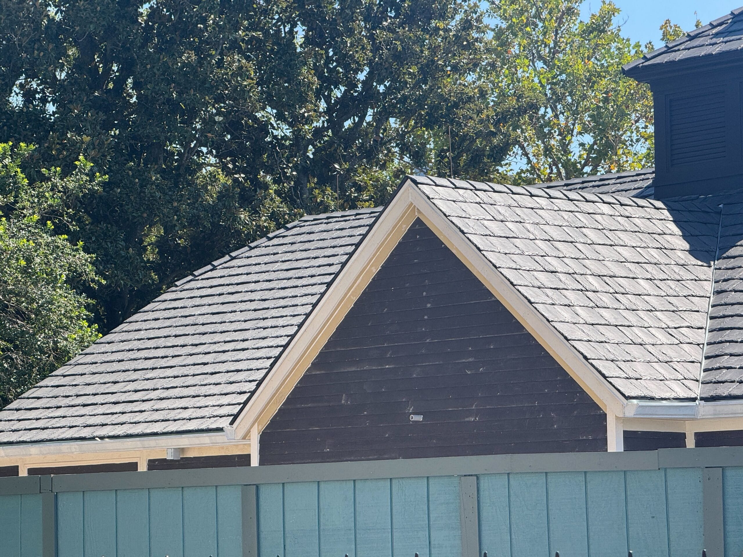 A dark brown house with a gray shingle roof behind a light blue wooden fence, trees in the background.