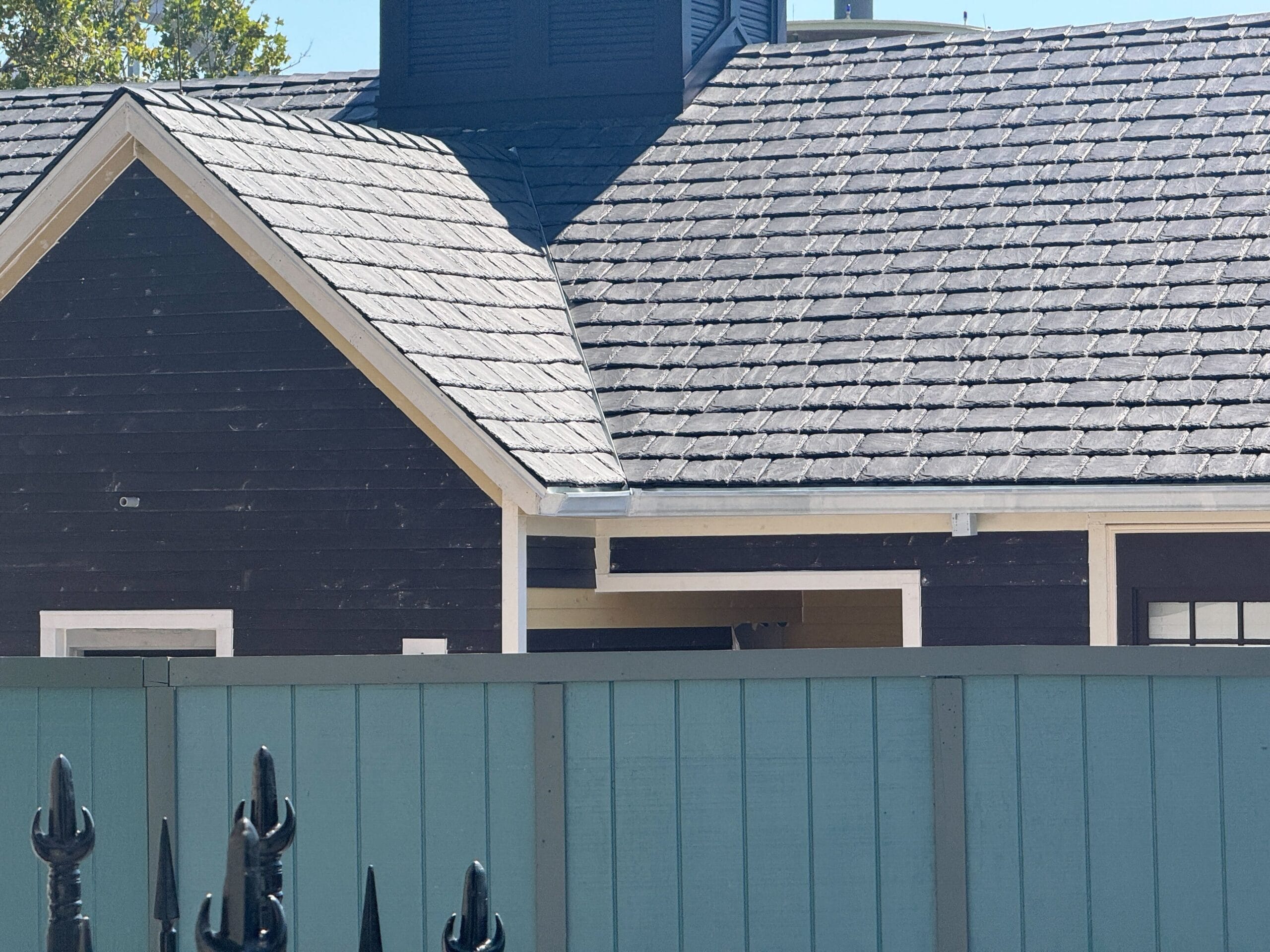 A dark wooden house with a shingled roof and a blue fence in front, seen on a sunny day.