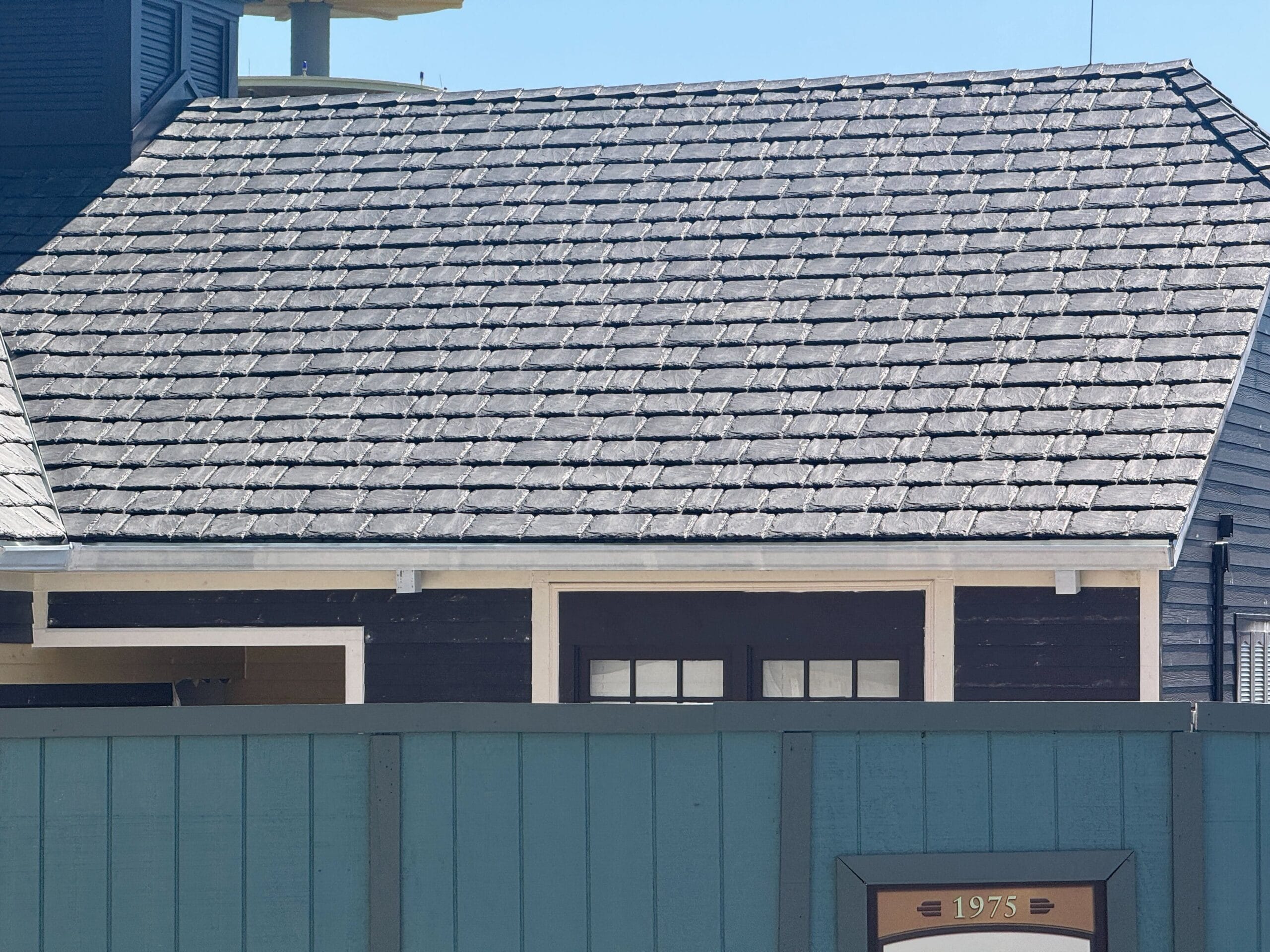 Dark shingled roof and upper wall of a building, with a teal fence and a sign reading "1975" in front.