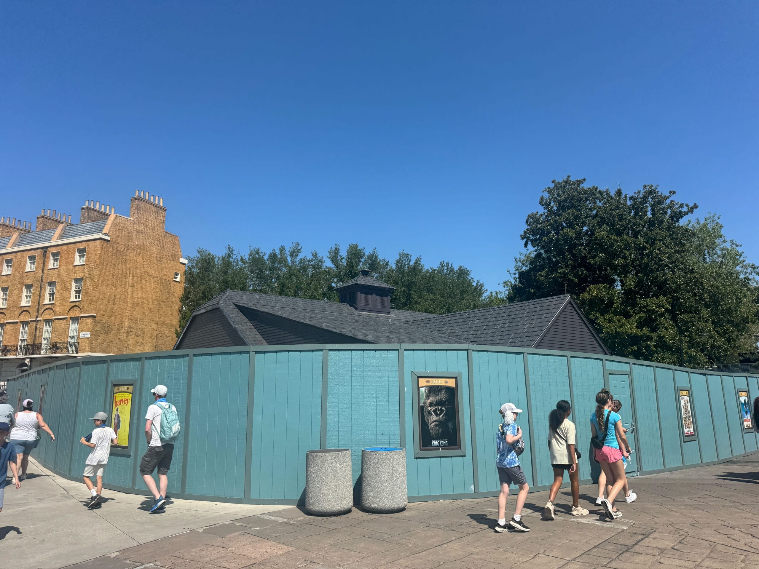People walk past a blue construction fence with movie posters on it, under a clear blue sky.