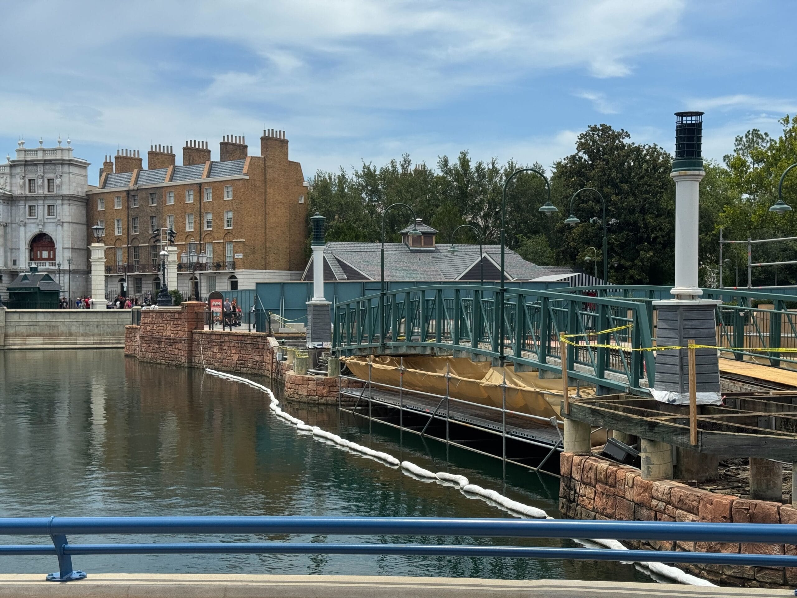 A green footbridge under construction over water, with buildings and trees in the background on a partly cloudy day.