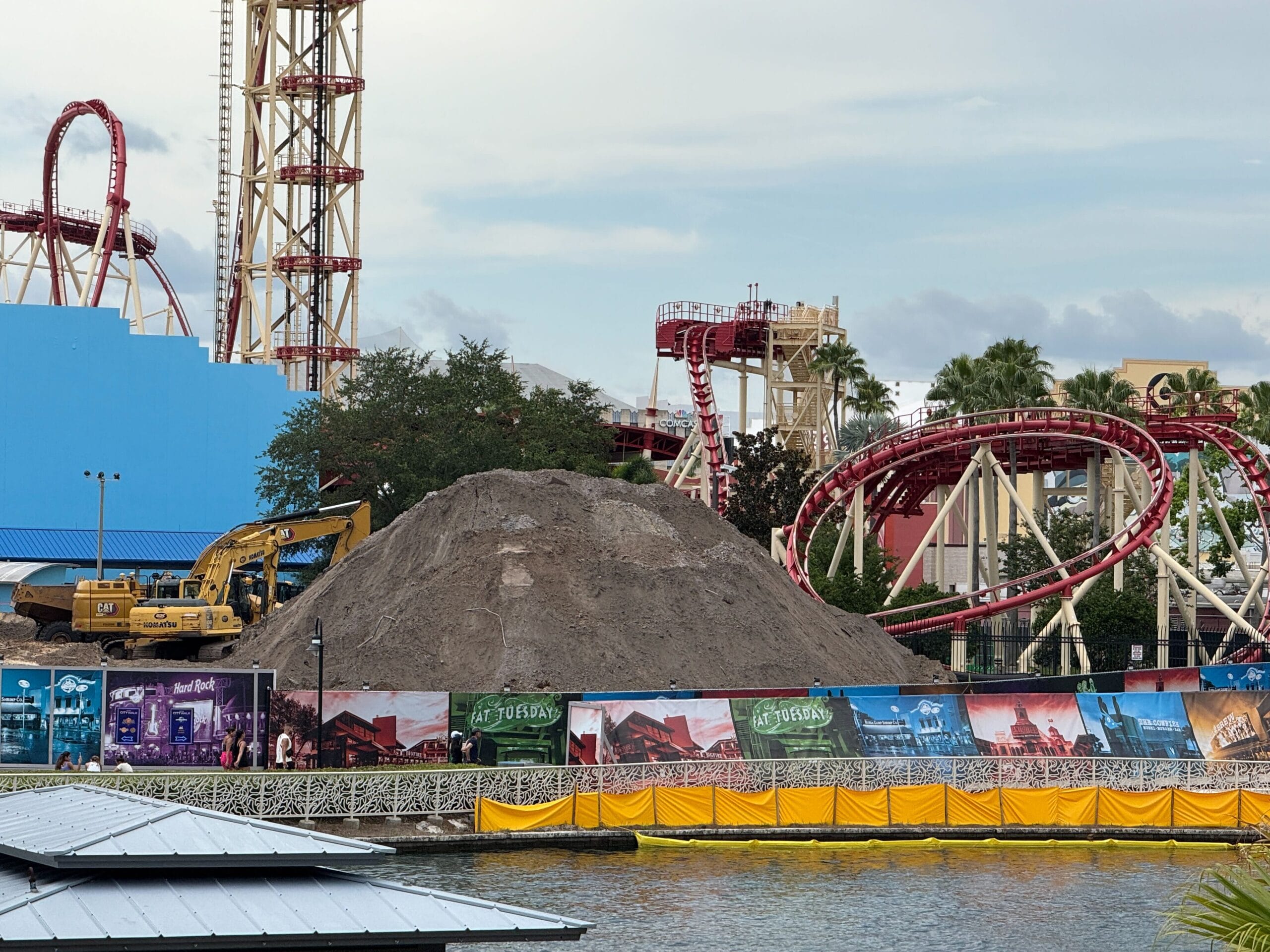 A large dirt mound and construction equipment sit in front of a red roller coaster at an amusement park.
