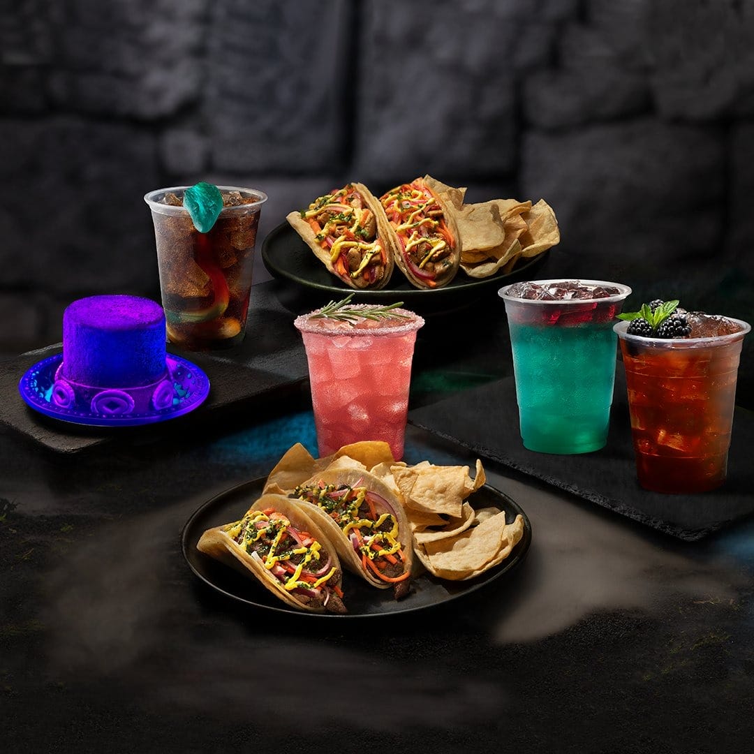 Three colorful drinks, tacos with chips, and a glowing purple hat set the scene for Halloween Horror Nights 2025.