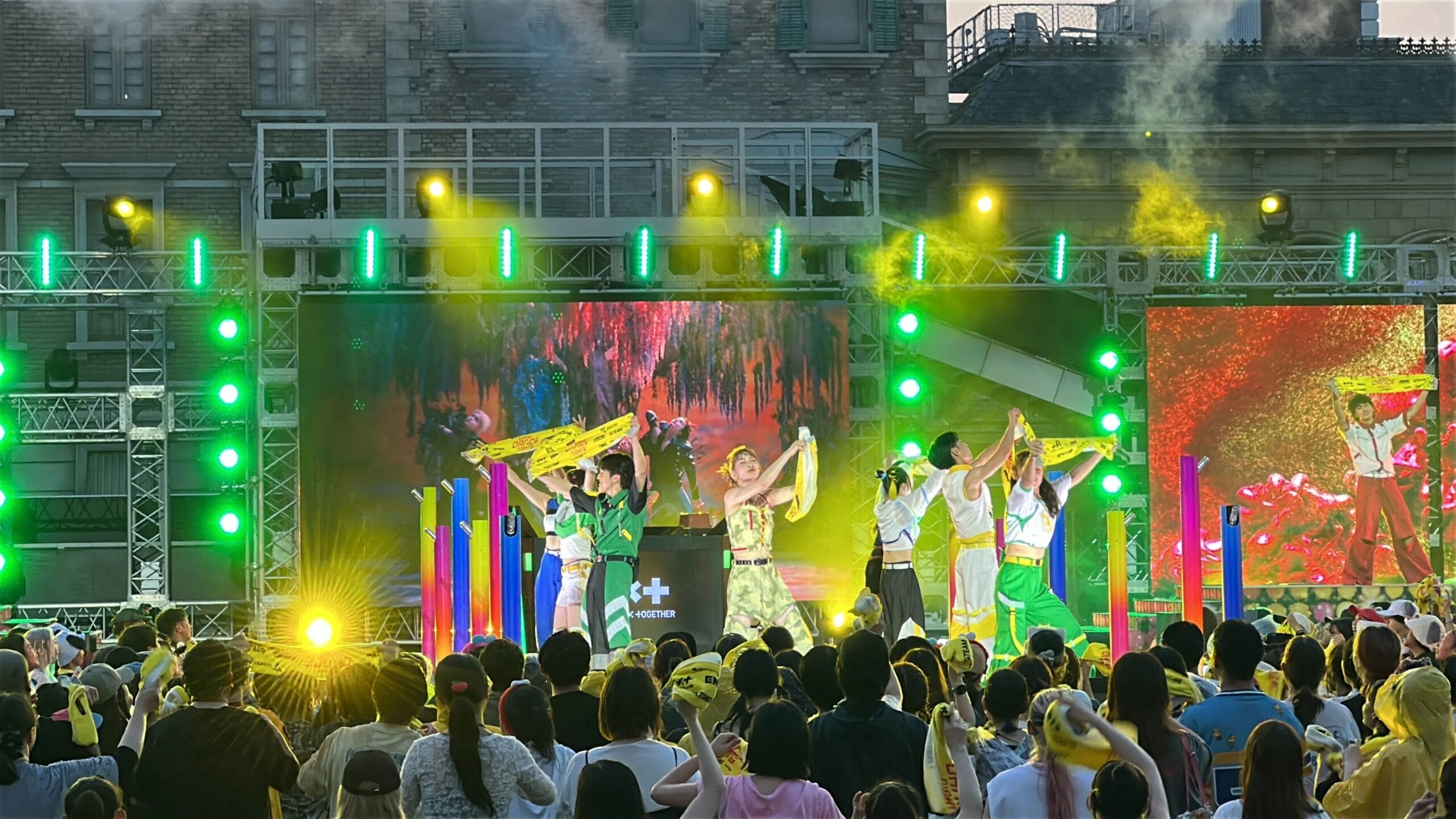 Performers in colorful costumes dance with yellow scarves at Summer Dance Nights, as crowds watch and capture photos.