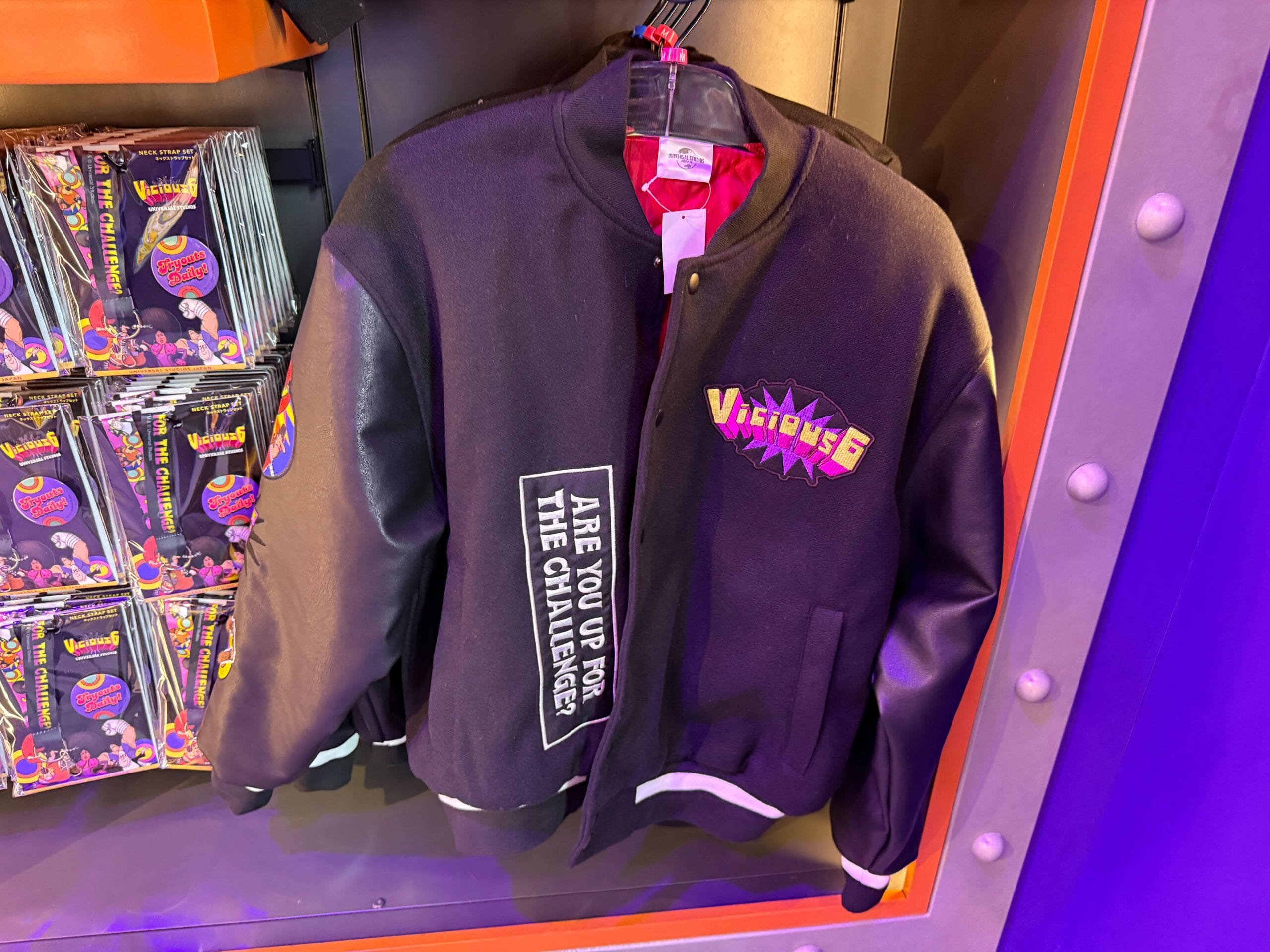 Black varsity jacket with "Villains 6" and "Are you up for the challenge?" patches, hanging in a store display.