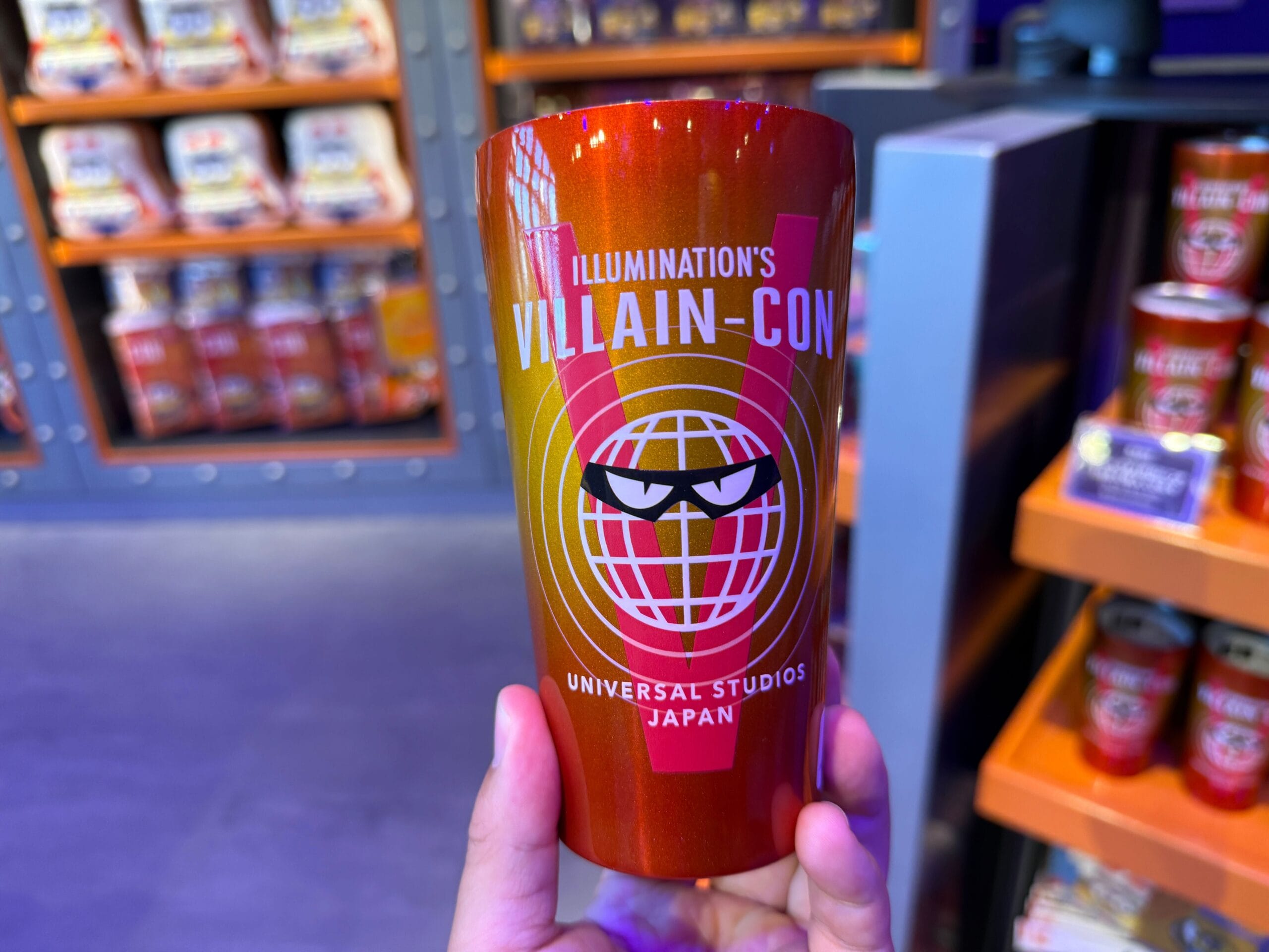 A hand holds a red Villain-Con cup from Universal Studios Japan, with a stylized globe and villain face design.