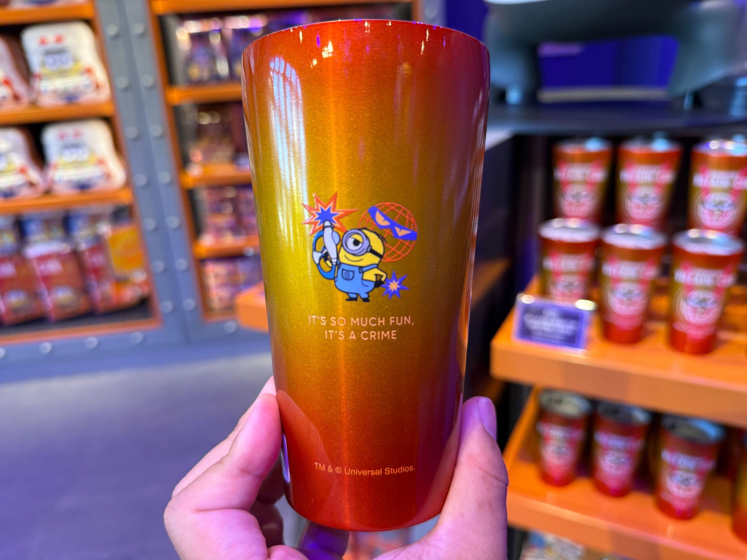 A hand holds a shiny red-orange Minions cup with text: "It's so much fun, it's a crime." Shelves are in the background.