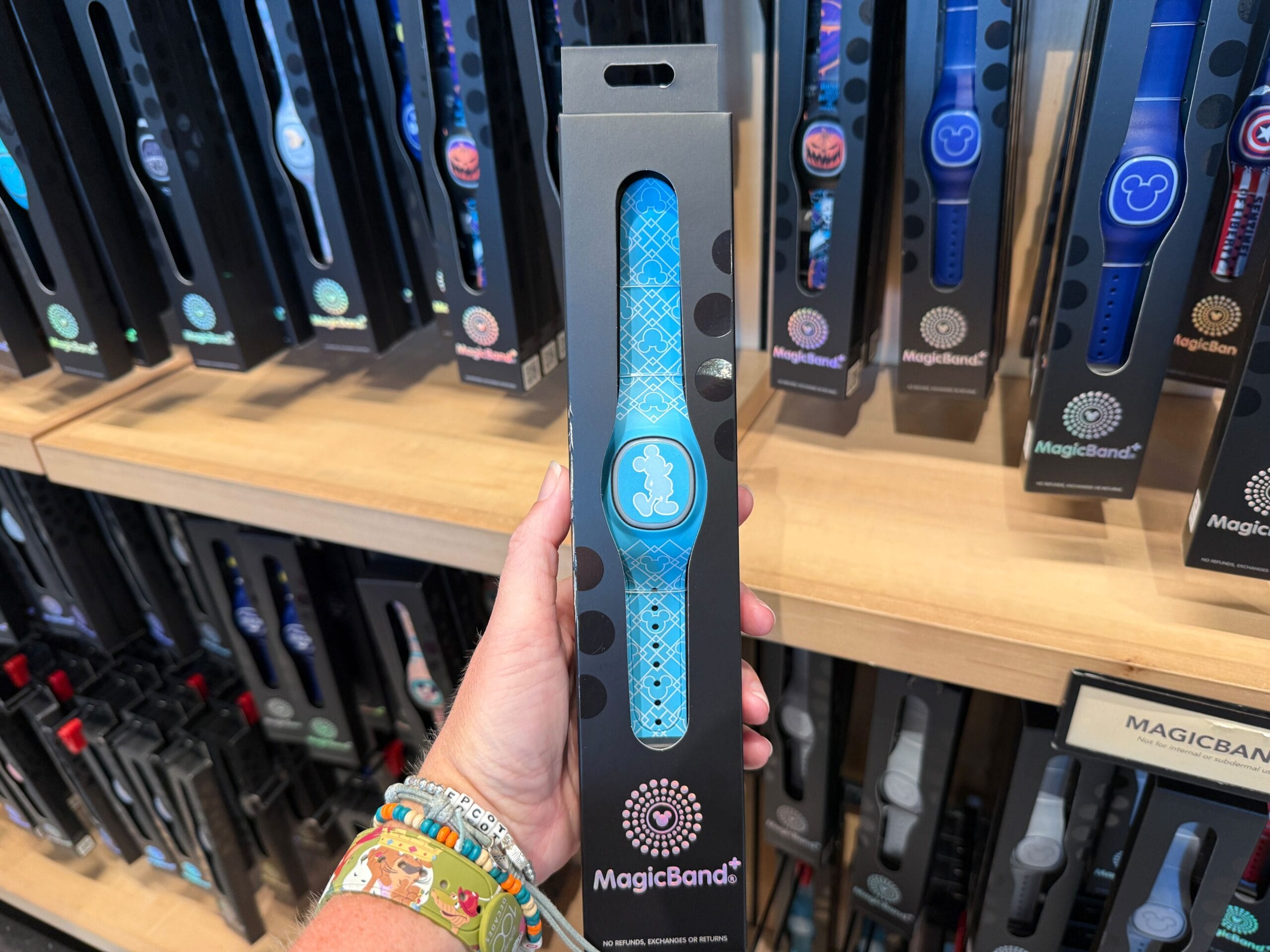 A hand holds a blue Disney MagicBand+ with a Mickey Mouse design in a store display with other bands.