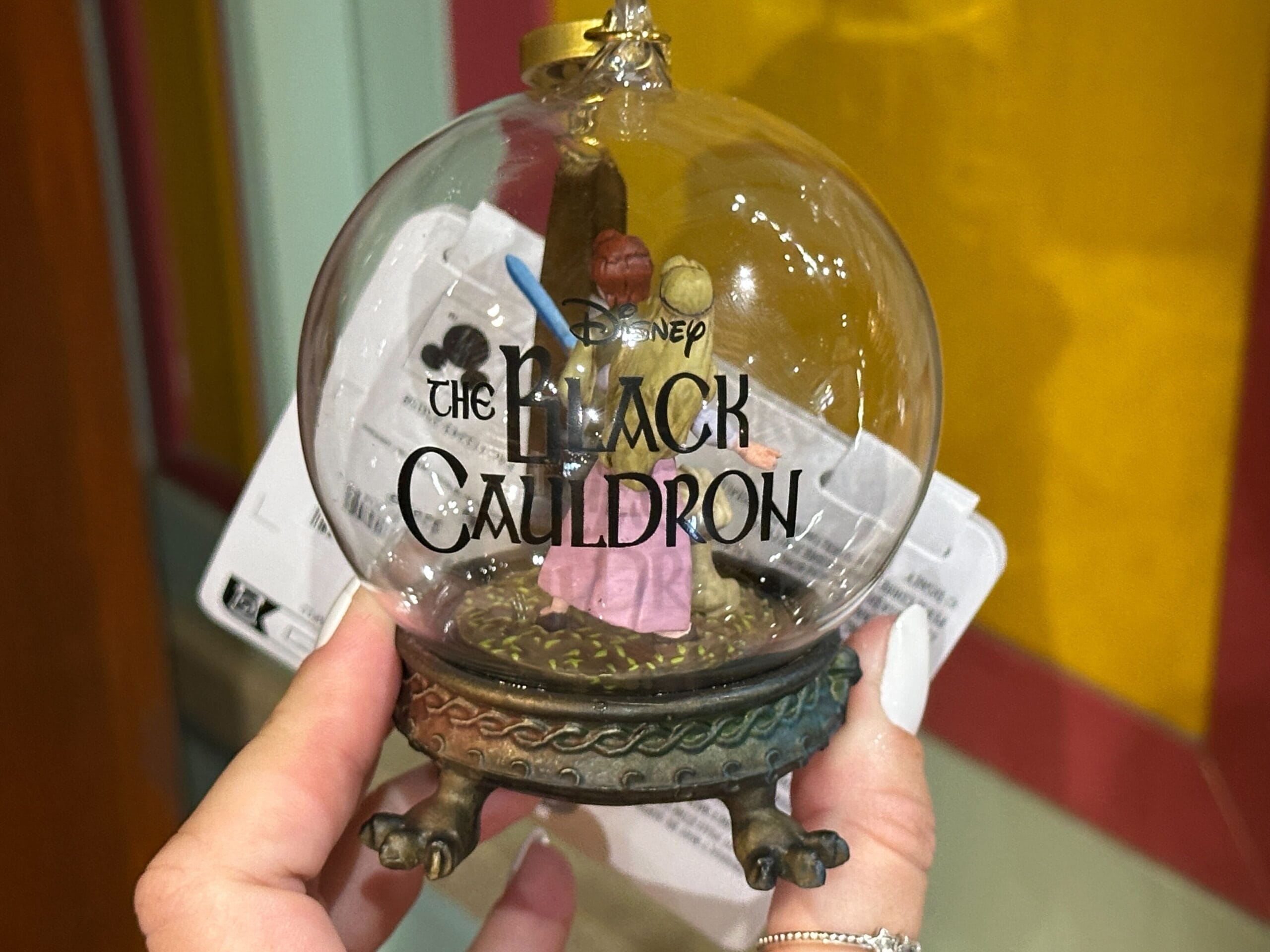 A hand holds a Disney “The Black Cauldron” snow globe ornament with two miniature figures inside.