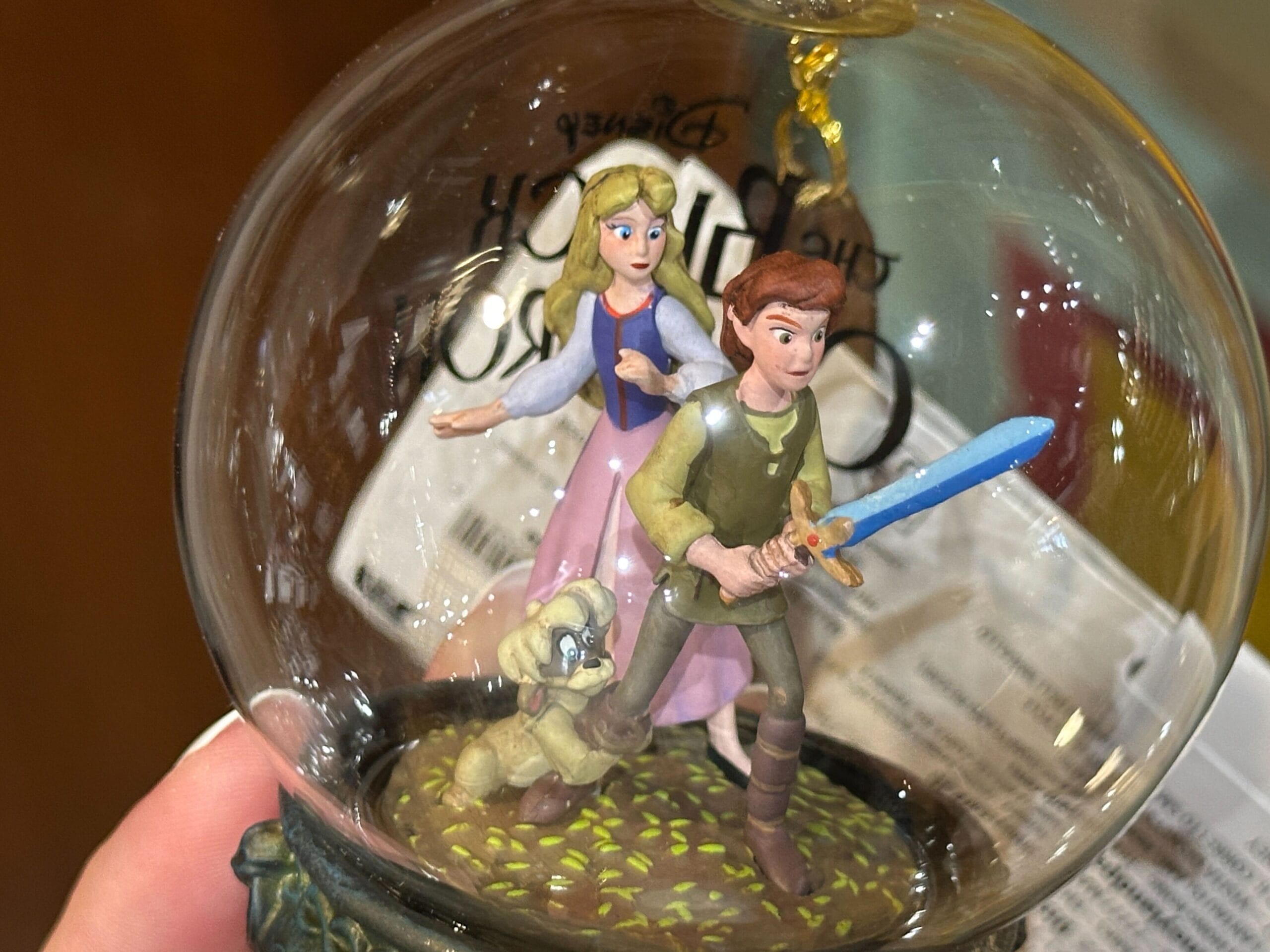 A hand holds a glass ornament featuring three fantasy characters, one with a sword, inside a dome.