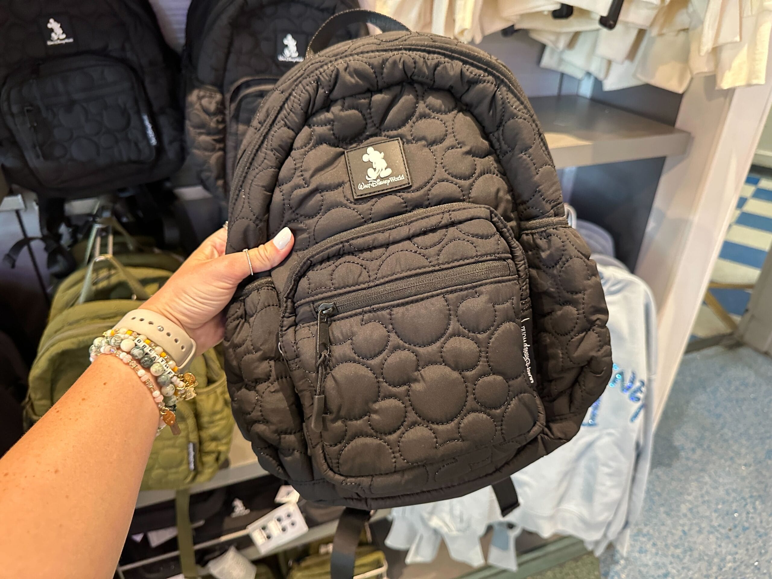 A hand holds a black quilted Ear Holder Backpack with a Mickey Mouse patch in a store display.
