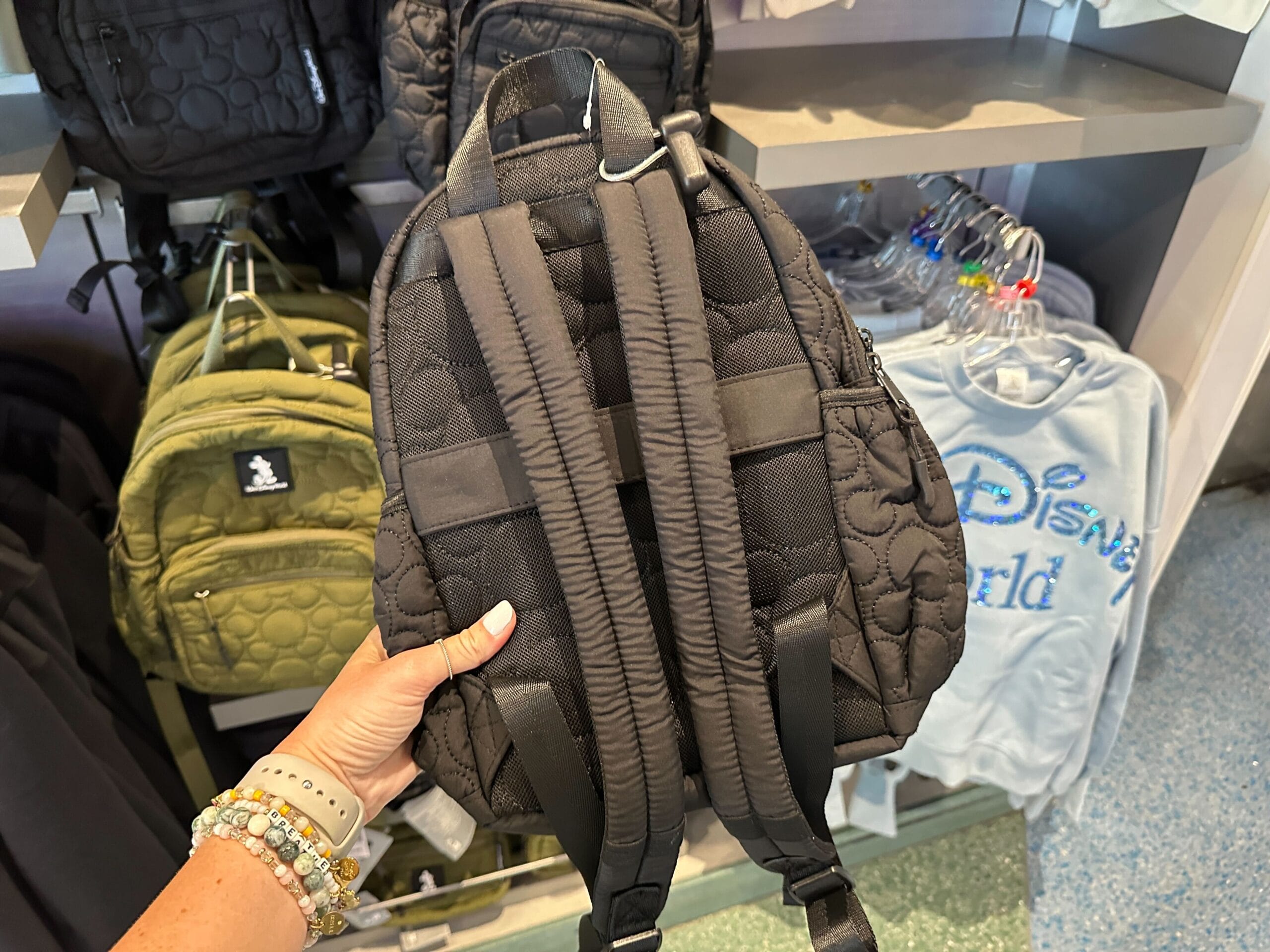 A hand holds a black Ear Holder Backpack in a store, with other bags and a blue Disney World sweatshirt in the background.