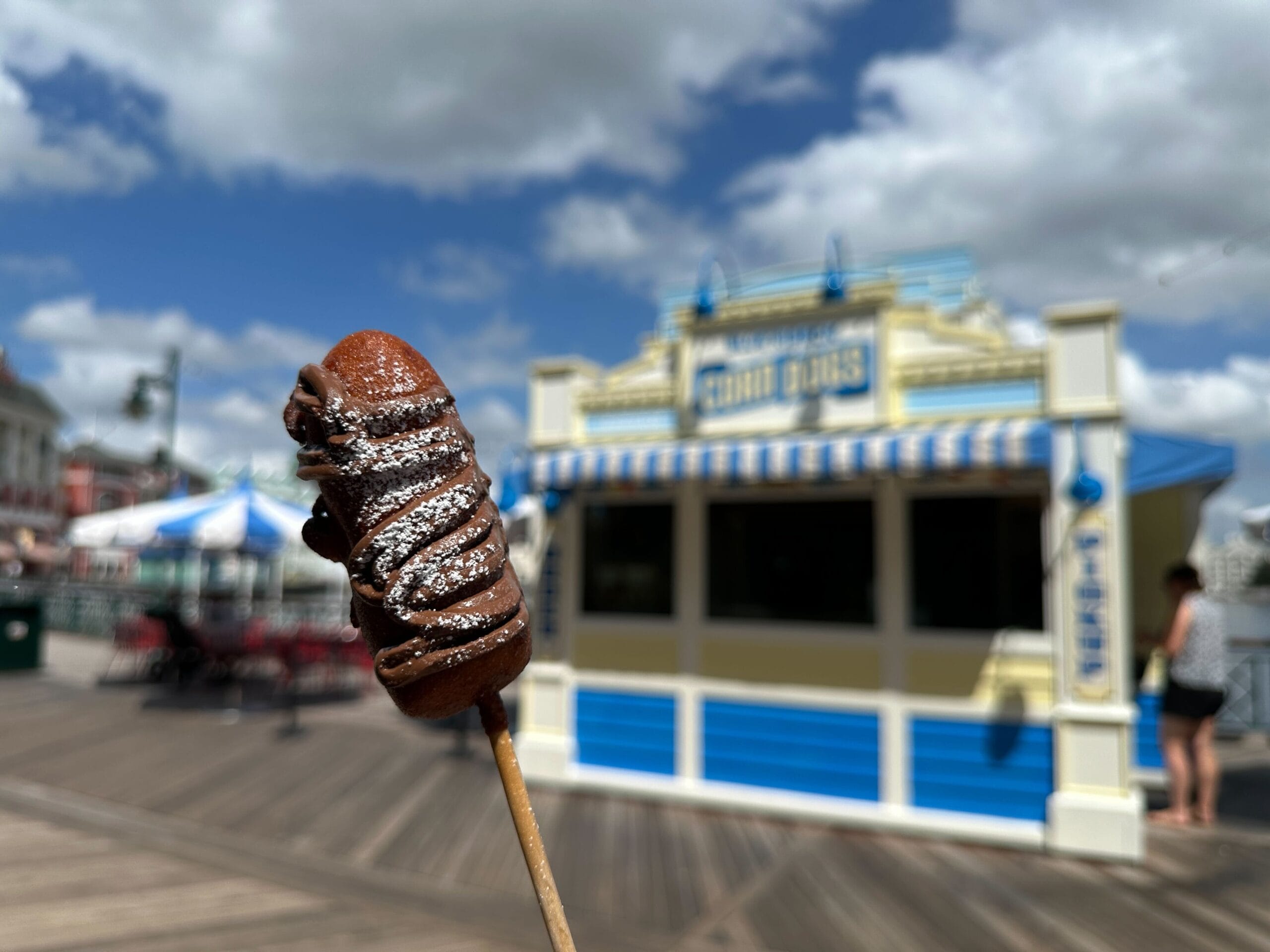 A chocolate-covered treat on a stick in front of a boardwalk food stand under a partly cloudy sky.