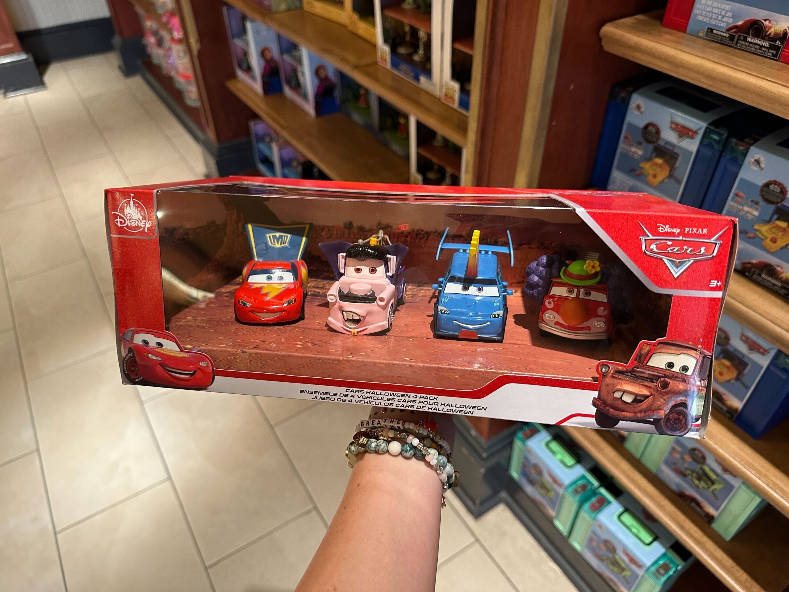 A hand holds a box with four Disney Pixar Cars toy cars on a store shelf. The Cars are all dressed in Halloween costumes.