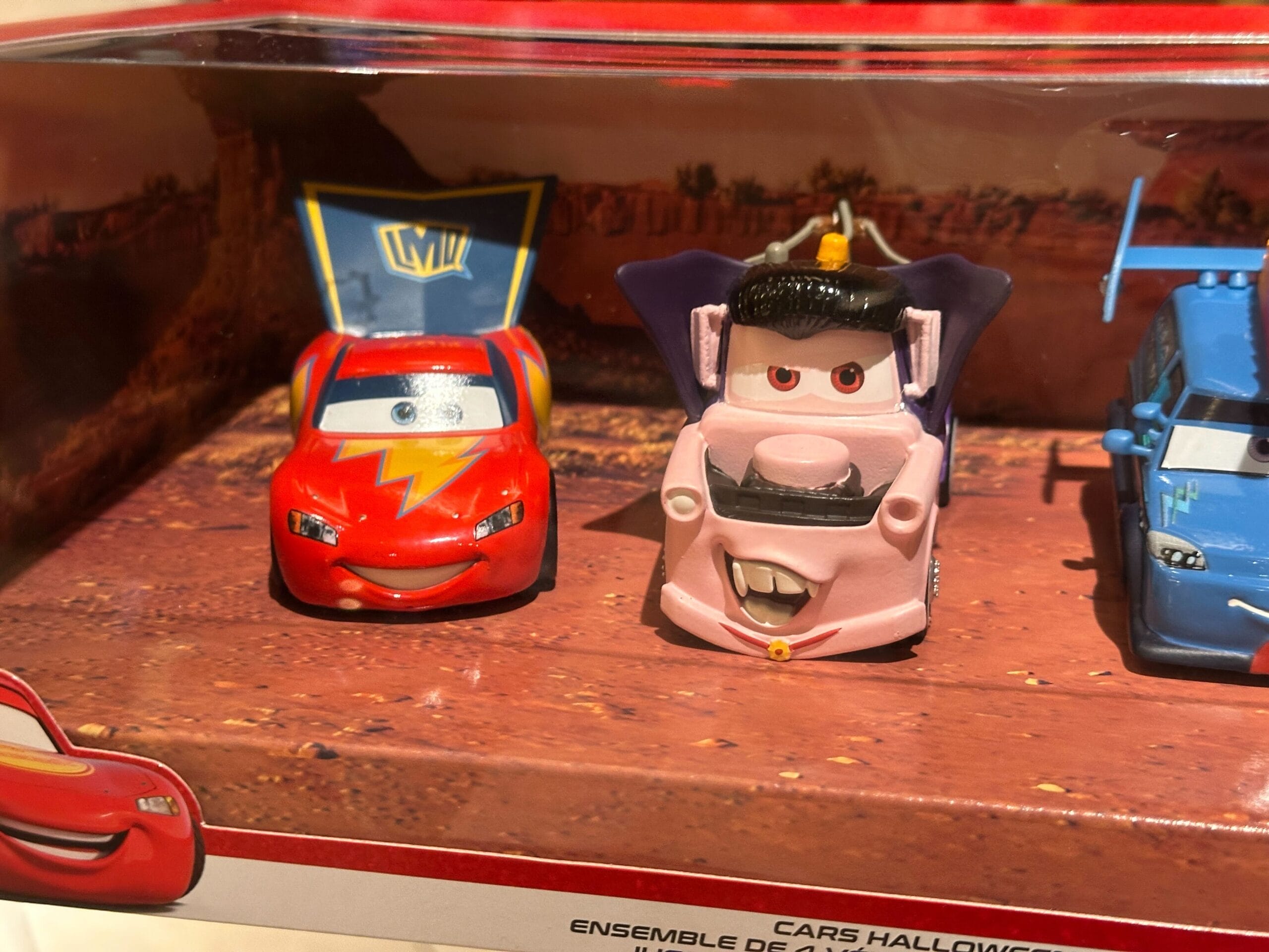 Toy cars from Cars, including one of Mater dressed as a vampire, displayed in a box with a desert background.